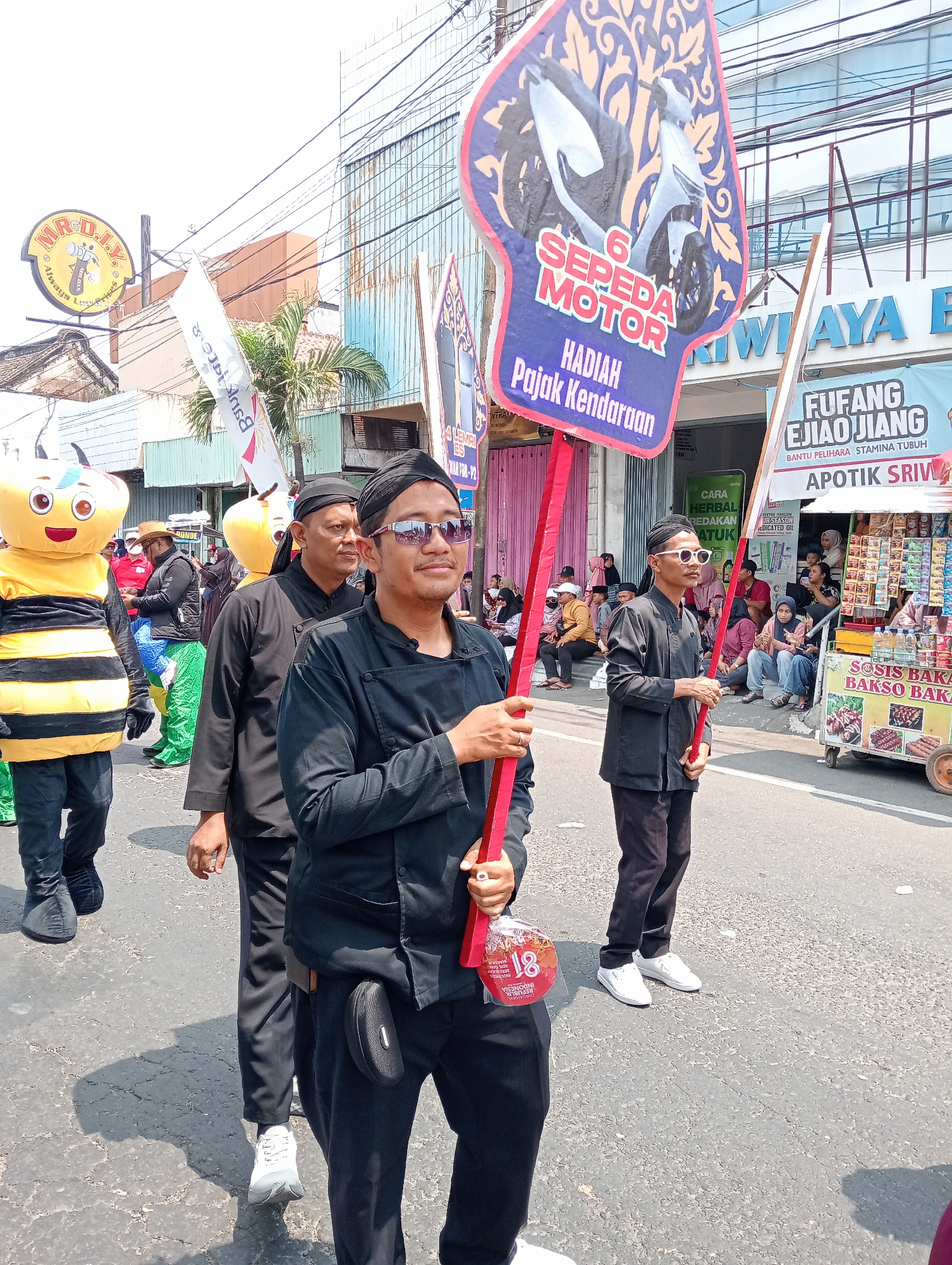 The image depicts a street scene in Demak, Indonesia, likely during a parade or festive event. In the foreground, several men are walking in formation, dressed in black attire and head coverings. One man in the center wears sunglasses and holds a large, elaborately decorated placard. This placard features an image of a motorcycle and text in Indonesian that translates to "6 Motorcycles, Prize, Vehicle Tax." To the left, a person in a bee costume with yellow and black stripes is also participating in the event. In the background, a building with blue siding is visible, along with banners and signs for local businesses, including "APOTIK SRIWIJAY" and "RUQANG EIJIAOJIANG." Spectators are seated on the side of the street, observing the procession. The ground is paved with asphalt, and sunlight indicates a daytime setting.