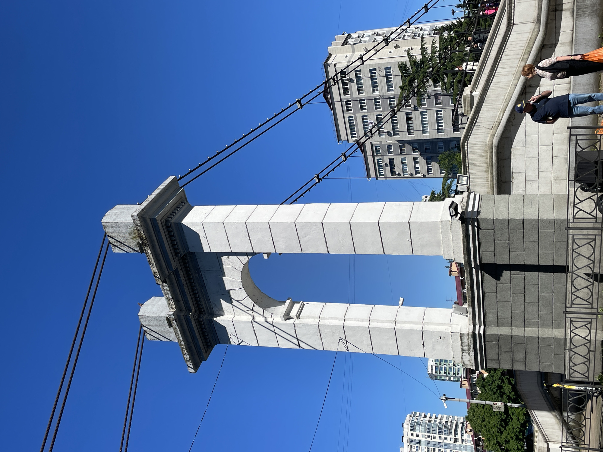 The image presents a bright, sunlit urban scene in Sochi, Russia, dominated by a large, light-colored concrete or stone architectural structure. This prominent feature, likely a bridge support or an ornate archway, consists of two tall, rectangular pillars connected by a wide arch at the top. Decorative molding adorns the structure near its upper and lower sections.
Several dark cables, possibly suspension cables or utility lines, crisscross the clear blue sky, some equipped with small, bright circular elements, resembling lights or insulators. Behind these cables and the main structure, a tall, multi-story building with a grey facade and numerous windows is visible. Another white multi-story building can be seen further in the background to the right.
On a stone walkway or platform to the right of the main structure, two individuals are present. One person, wearing a dark blue shirt, light blue jeans, and a dark baseball cap, stands facing forward. Slightly behind them is another person, possibly female, with reddish-brown hair, dressed in a patterned top and carrying an orange bag. A group of other individuals is partially visible further back, near a lush green evergreen tree. An ornate black metal railing lines the edge of the walkway.
The time of day appears to be bright daytime, with a clear, cloudless blue sky and strong sunlight casting distinct shadows from the architecture and people. The setting conveys a vibrant, sunny urban atmosphere. No discernible text is visible within the image.