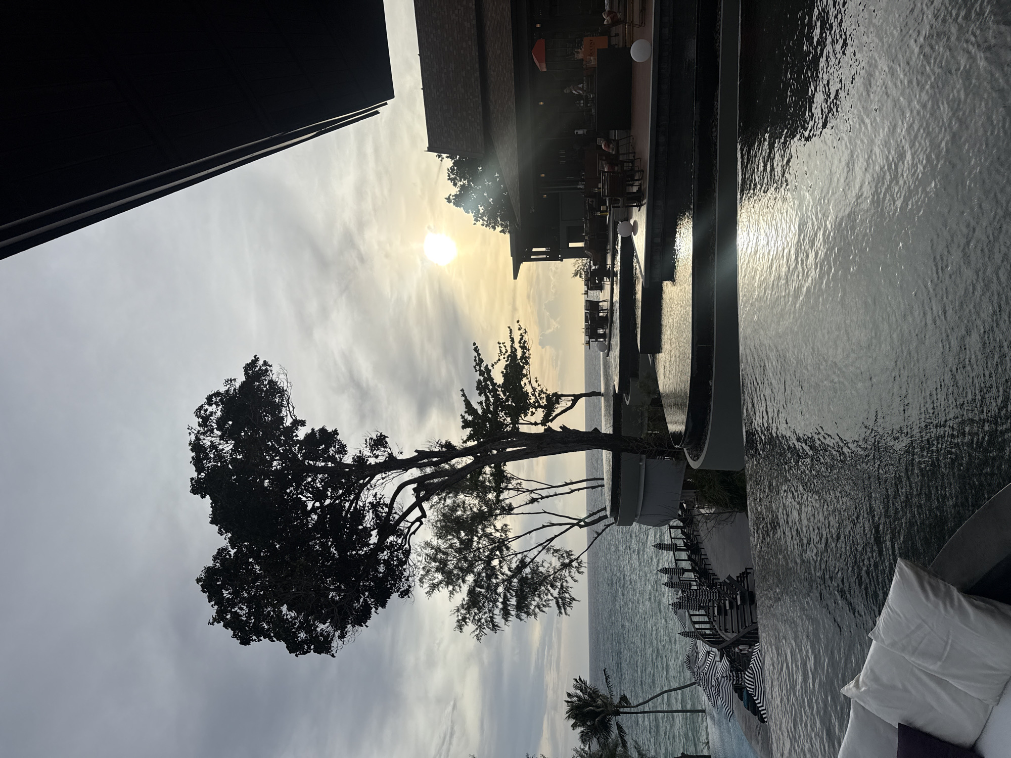 The image depicts a resort in Sakhu, Thailand, at dusk. A body of water, possibly a swimming pool or infinity pool, dominates the foreground, reflecting the muted light of the setting sun. Behind the water, a multi-tiered structure with several pools is visible, with tables and chairs set up for dining. The sea stretches out to the horizon, with a few palm trees and other vegetation lining the shore. A large, dark tree with a distinctive heart-shaped canopy stands prominently in the mid-ground. A building with a dark, sloping roof is visible to the right, with people seated at tables under an orange umbrella. The sky is overcast with a bright sun peeking through the clouds. Beach umbrellas with black and white stripes are arranged along a walkway bordering the sea. The overall atmosphere is serene and luxurious.