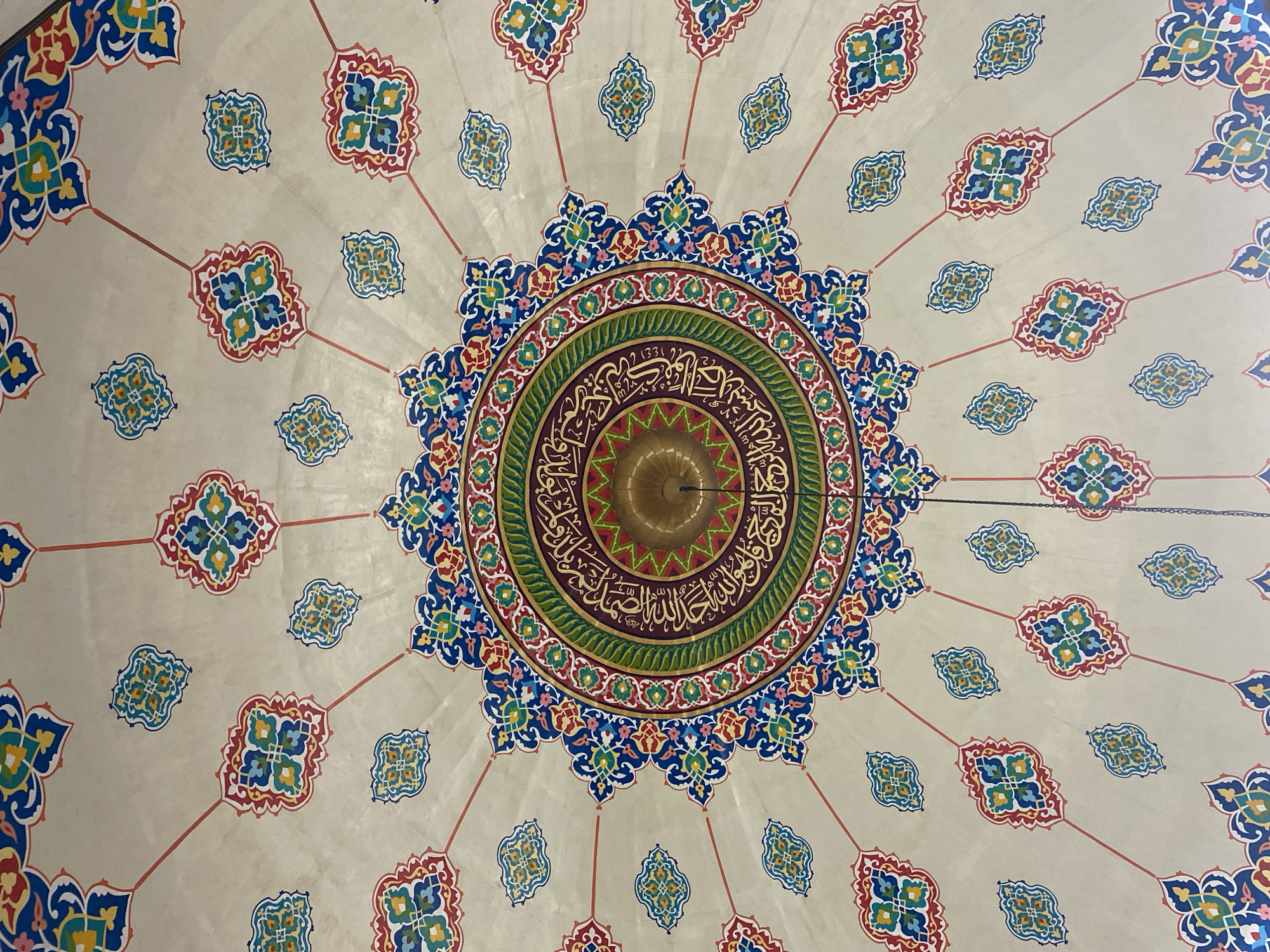 The image displays the ornate dome of the Islamic Center Mosque in Lhokseumawe, Indonesia. The dome features intricate geometric patterns and Arabic calligraphy painted in vibrant colors, including blue, red, green, yellow, and gold, set against a light beige background. The central area of the dome is dominated by a circular design with concentric rings of decoration, culminating in a gilded finial at its apex. Radiating from the center are delicate red lines, interspersed with repeating motifs of stylized floral or paisley shapes in various colors. These smaller decorative elements are arranged radially across the dome's surface. A thin chain hangs down from the finial, partially visible in the lower right quadrant of the image. The overall impression is one of rich Islamic artistry and architectural detail.