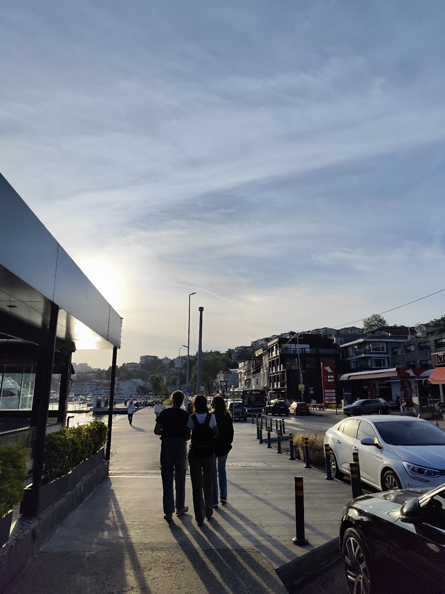 The image depicts a late afternoon or early evening scene along a promenade in Sarıyer, Turkey. The sun is low on the horizon, positioned to the left, casting long shadows across the concrete walkway and illuminating the sky with a warm glow.
In the foreground, three people are walking away from the camera, their figures silhouetted against the sunlight, casting distinct, elongated shadows. To their left, a sleek, modern building with a dark, reflective facade runs alongside the promenade. Beyond this building, a body of water, likely the Bosphorus, is visible with several boats and yachts moored.
Parallel to the promenade on the right, a road is present with cars parked and some moving traffic. Black and yellow bollards delineate the pedestrian area from the street. On the far side of the road, various buildings climb a gentle hillside. A prominent feature is a gas station, partially visible, displaying "62.52" and "65.27" on its pricing sign, and the brand "MAX". The sky is a clear light blue with thin, wispy clouds, and a subtle contrail is visible high above. The scene captures a peaceful urban waterfront setting.