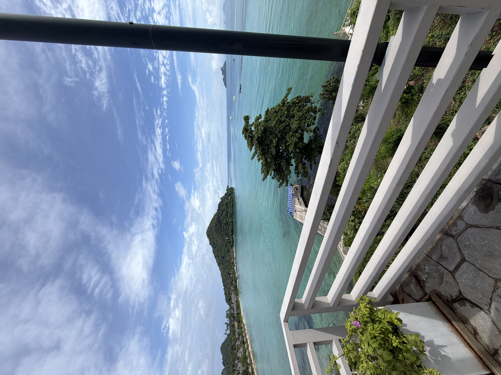 A serene view overlooks a turquoise bay from a balcony in Ao Nang, Thailand. The foreground features a white railing and a potted plant with pink flowers. Beyond the railing, a stone-tiled patio leads to the edge of the balcony. The bay is dotted with several boats, and a narrow pier with blue and white checkered coverings extends into the water. A lush green tree stands on the right side of the pier. In the distance, a verdant, mountainous island with a sandy beach and resort buildings lines the bay. The sky is bright blue with scattered white clouds.