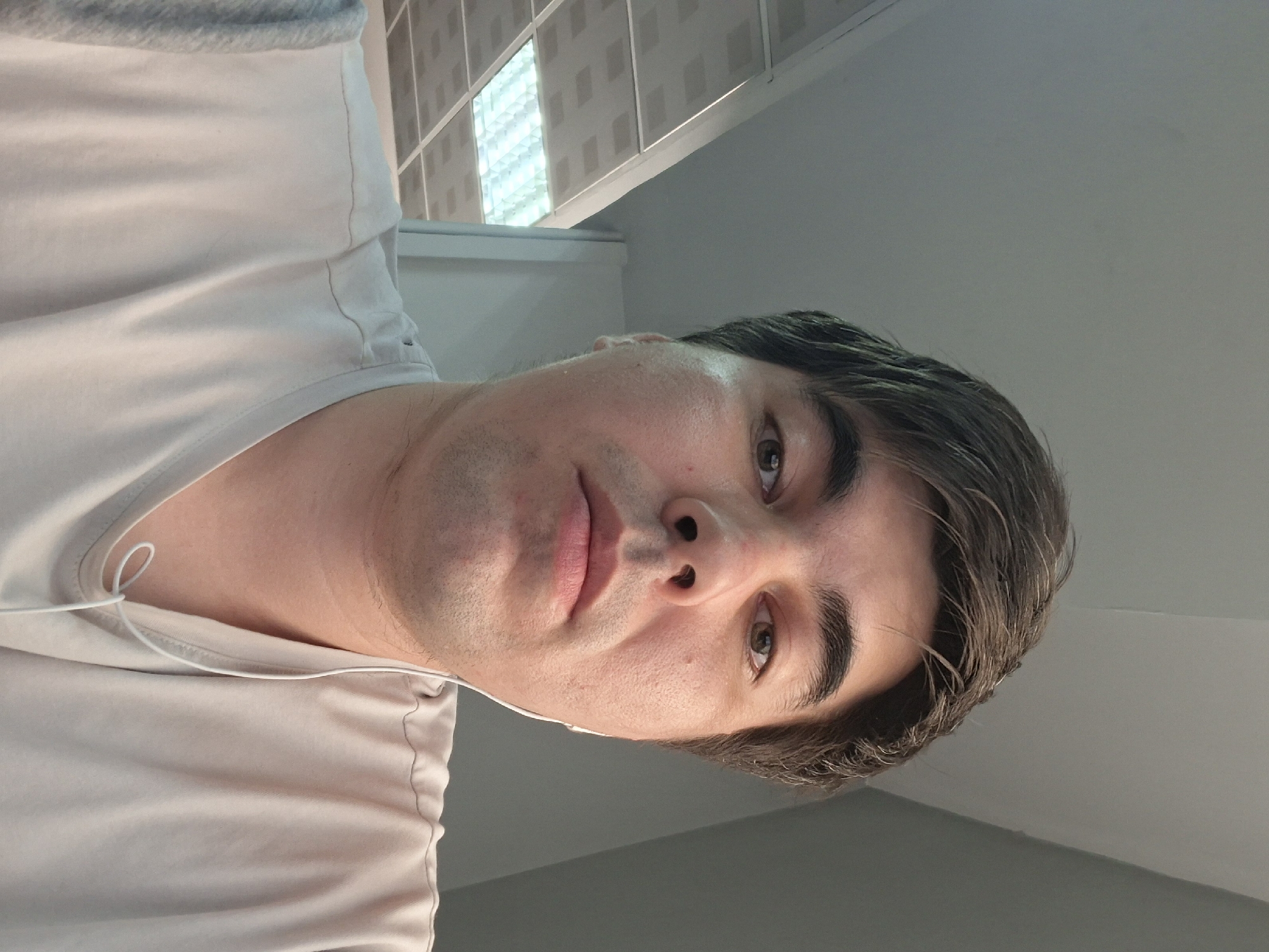 The image is a selfie of a man taken from the chest up. He has light skin, dark brown hair, and brown eyes. He is wearing a light-colored t-shirt and white earbuds. The background appears to be an indoor environment, possibly an office or a public space, with a ceiling made of fluorescent light panels. The man's facial expression is neutral, and he is looking directly at the camera. There is no specific indication of his location within the city of Astana, Kazakhstan, in the image itself.