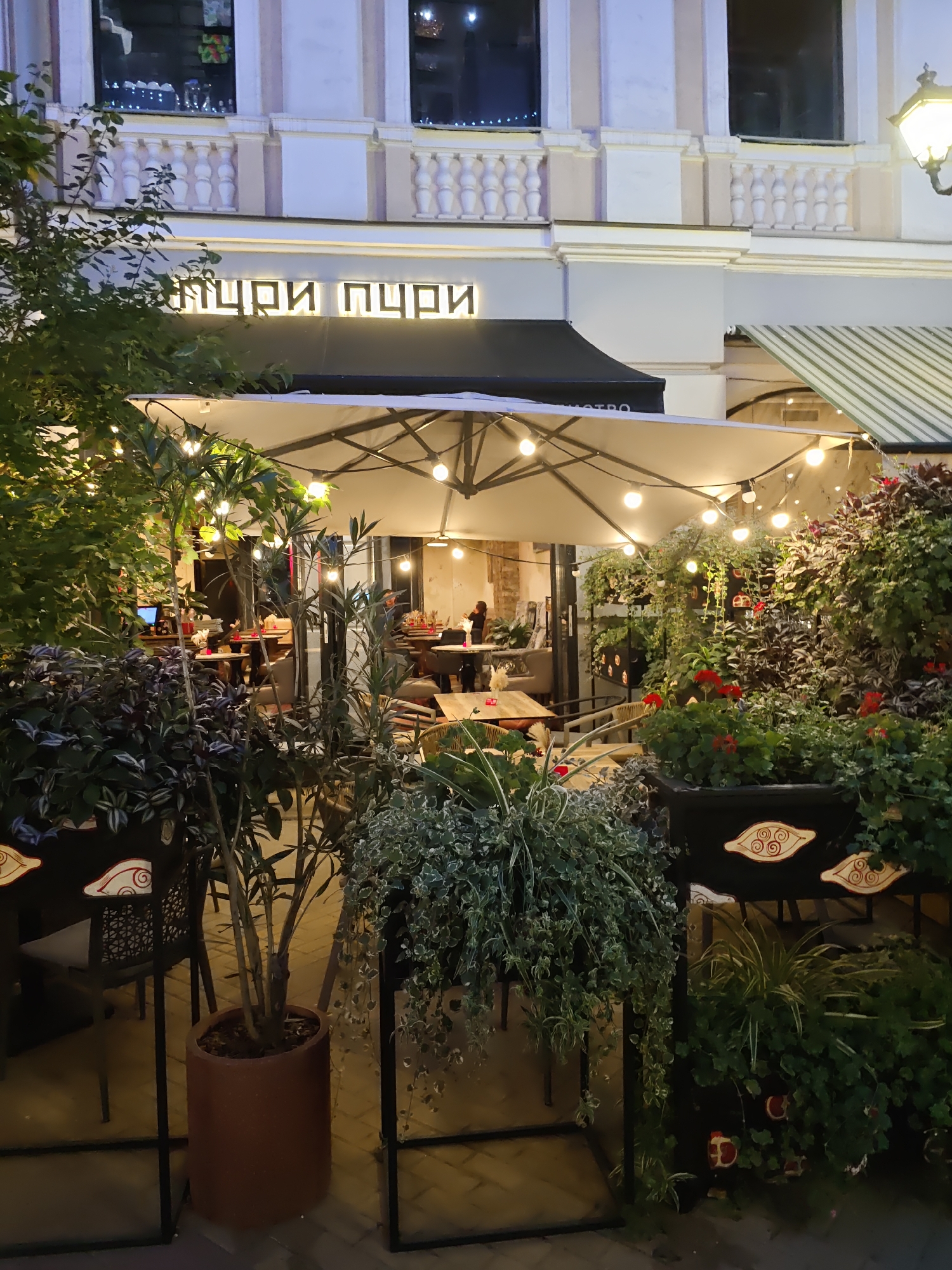The image shows the exterior of a restaurant in Rostov-on-Don, Russia. The restaurant features an outdoor seating area with tables and chairs under a large umbrella with string lights. There are several potted plants and greenery surrounding the seating area, creating a garden-like atmosphere. The building facade above the restaurant has classical architectural details, including columns and balustrades, and is illuminated at night. A sign with Cyrillic lettering is visible above the awning. The overall scene suggests a cozy and inviting dining establishment during evening hours.