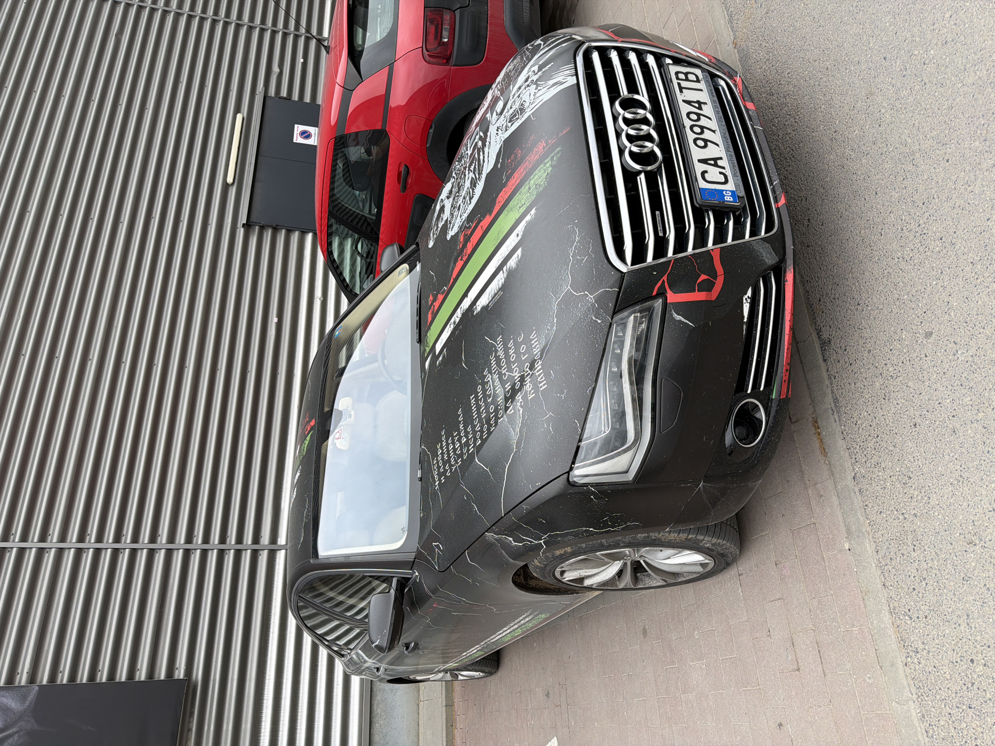 The image shows the front of a black Audi car parked on a paved area, likely a street or parking lot. The car has distinctive custom artwork or a wrap featuring white scribbles, red and green stripes, and what appears to be script in a foreign language. The car's Bulgarian license plate reads "CA 9994 TB" and includes a small BG flag. Behind the Audi, partially visible, is a red car, possibly a hatchback, parked next to a corrugated metal wall with a grey rectangular panel on it. The text on the front of the Audi car references, "CD. Porsche Plovdiv" indicating that the car is in Plovdiv, Bulgaria. The area appears to be urban, likely an industrial or commercial area due to the type of building wall.
