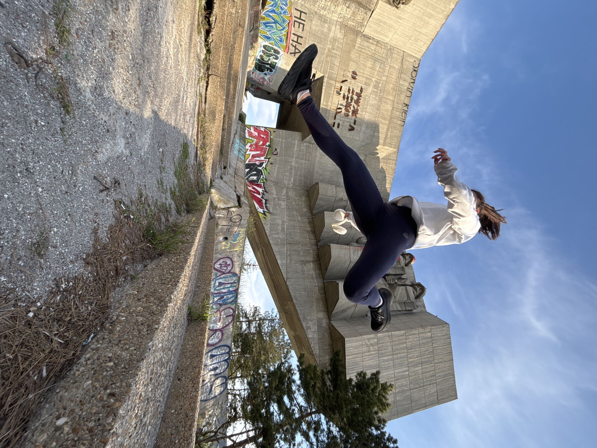 The image shows a person mid-air, likely jumping. The individual is wearing dark leggings, a white hooded sweatshirt, and dark shoes. The setting appears to be an urban location, specifically near a large concrete structure with graffiti. The backdrop includes a blue sky. The ground area consists of gravel and concrete. The location is Unknown, Unknown.