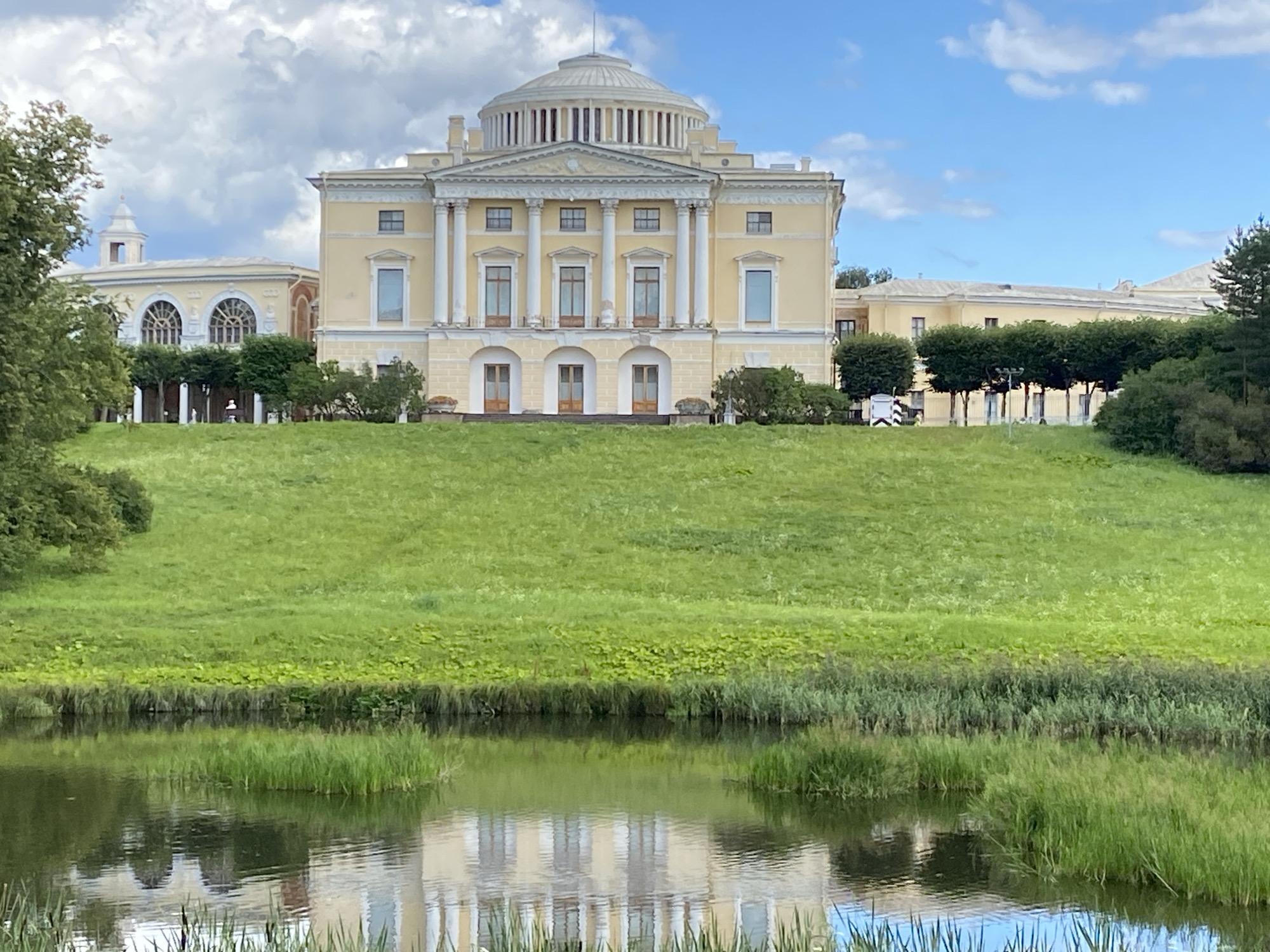 The image shows a grand, light yellow neoclassical palace with a prominent dome, located in the city of Pavlovsk, Russia. The palace is situated atop a lush green hill, which slopes down towards a reflective body of water in the foreground. The water mirrors the architecture of the palace, as well as the surrounding trees and the sky.
The palace features classical architectural elements such as large columns, arched doorways, and numerous windows. To the left of the main structure, a lower, connected wing with arched windows is visible. A row of manicured trees lines the base of the hill in front of the palace.
The environment is characterized by vibrant green grass covering the expansive hill and denser vegetation near the water's edge. The sky is blue with scattered white clouds. The overall scene depicts a well-maintained park or estate, bathed in natural light under a partly cloudy sky. There are no visible people or animals in the image.