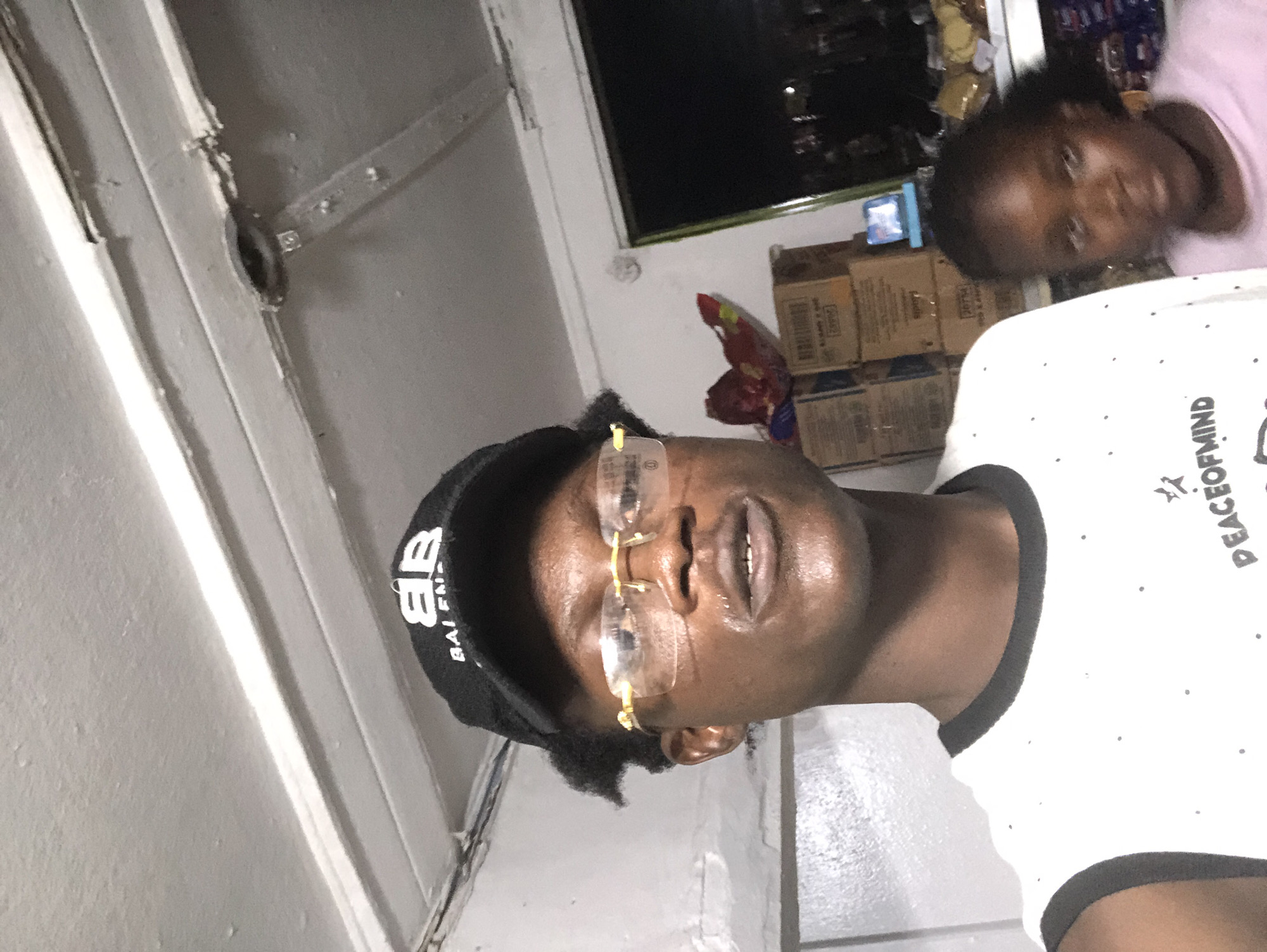 A young male, wearing a black baseball cap and gold-rimmed glasses, is captured in a selfie. He has short, dark hair and is wearing a white t-shirt with black trim. Behind him, a younger female with dark hair is visible, appearing to be in a store or warehouse. The background shows stacked cardboard boxes, a red and yellow item, and a dark opening that suggests an exterior view or a dimly lit area. The overall environment appears to be a commercial space, possibly a shop or storage facility, consistent with a location in Ado, Nigeria.