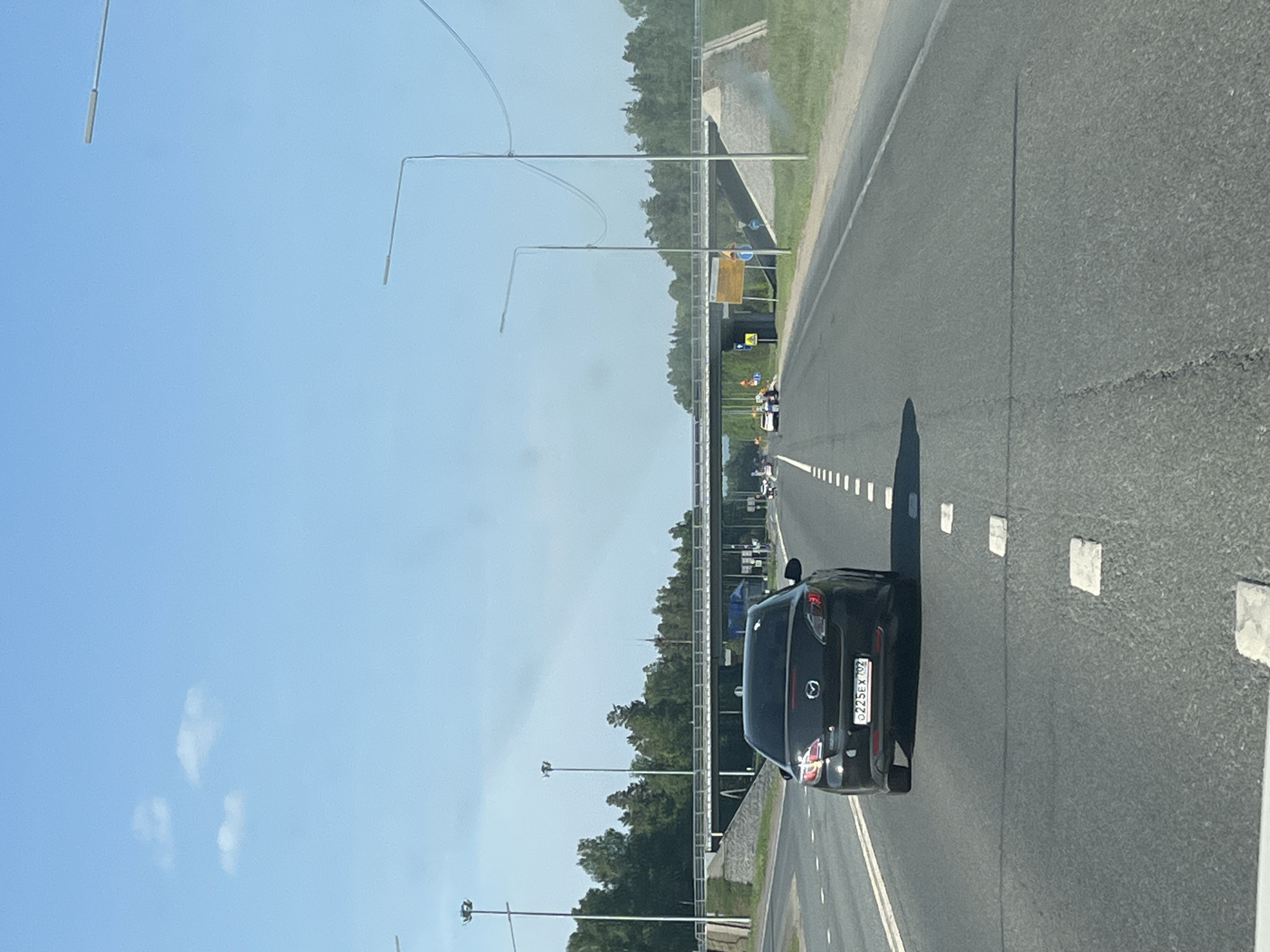 A photograph depicts a highway in what is presumed to be Severnyy, Russia, based on the vehicle license plate. A dark-colored Mazda is in the right lane of the highway. The scene is captured during daytime under a clear sky with sparse clouds. Streetlights line the side of the highway, which is surrounded by greenery. There are several vehicles in the distance. A bridge is visible over the highway. The license plate of the Mazda reads "c225ex 702".