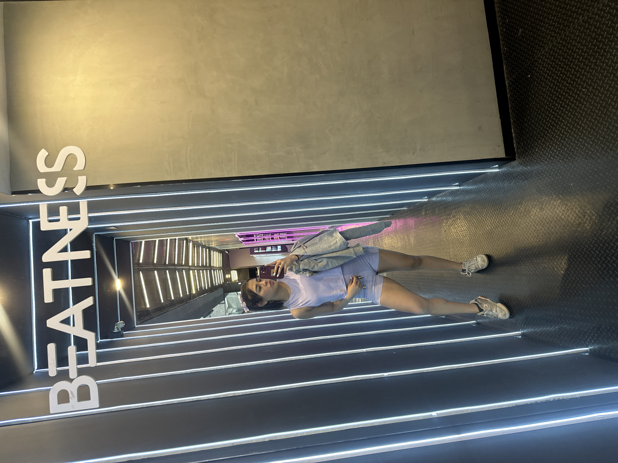 A young woman is centered in a long, narrow hallway with horizontally oriented light strips illuminating the black walls and floor. The floor has a textured, diamond-patterned surface. The text "BEATNESS" is displayed vertically in white on the left wall near the entrance of the hallway. The woman is wearing athletic apparel and sneakers. She is holding a phone, as if taking a selfie or video. A denim jacket is draped over her arm. The exact location in San Bernardino Tlaxcalancingo, Mexico cannot be determined from the provided information.
