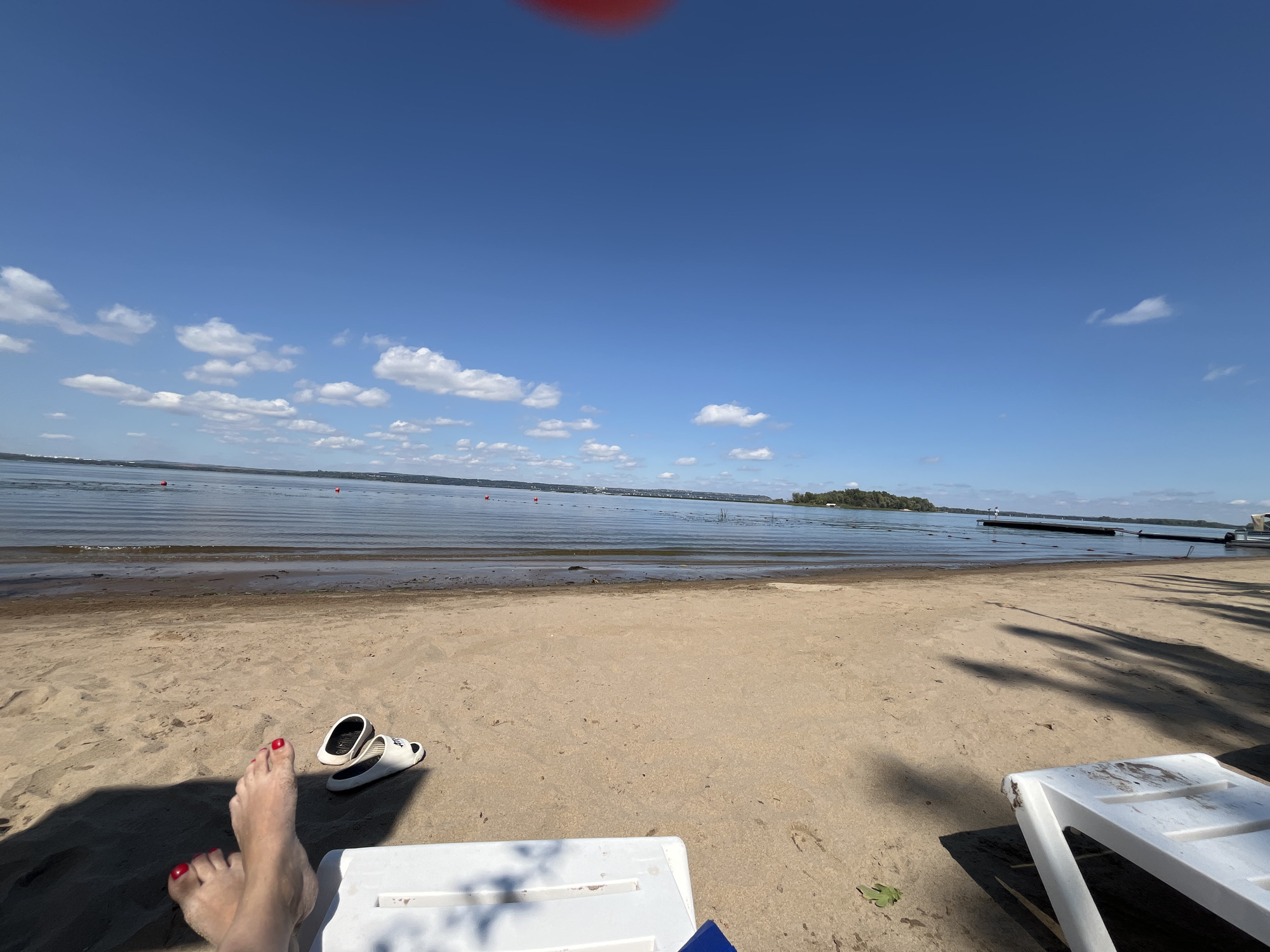 The image depicts a sandy beach on a sunny day. The foreground shows a pair of bare feet with red painted toenails, appearing relaxed and slightly covered in sand. Next to the feet is a pair of white and black sandals. A white beach chair, partially in shadow, is positioned in the lower foreground. Beyond the beach, a calm body of water stretches towards the horizon, with a faint shoreline visible in the distance. Several red buoys are scattered across the water, indicating a designated swimming area or boundary. The sky is a clear blue with scattered white clouds. The overall scene suggests a peaceful and leisurely day at a beach, possibly in the city of Shumeika, Russia, given the context provided.