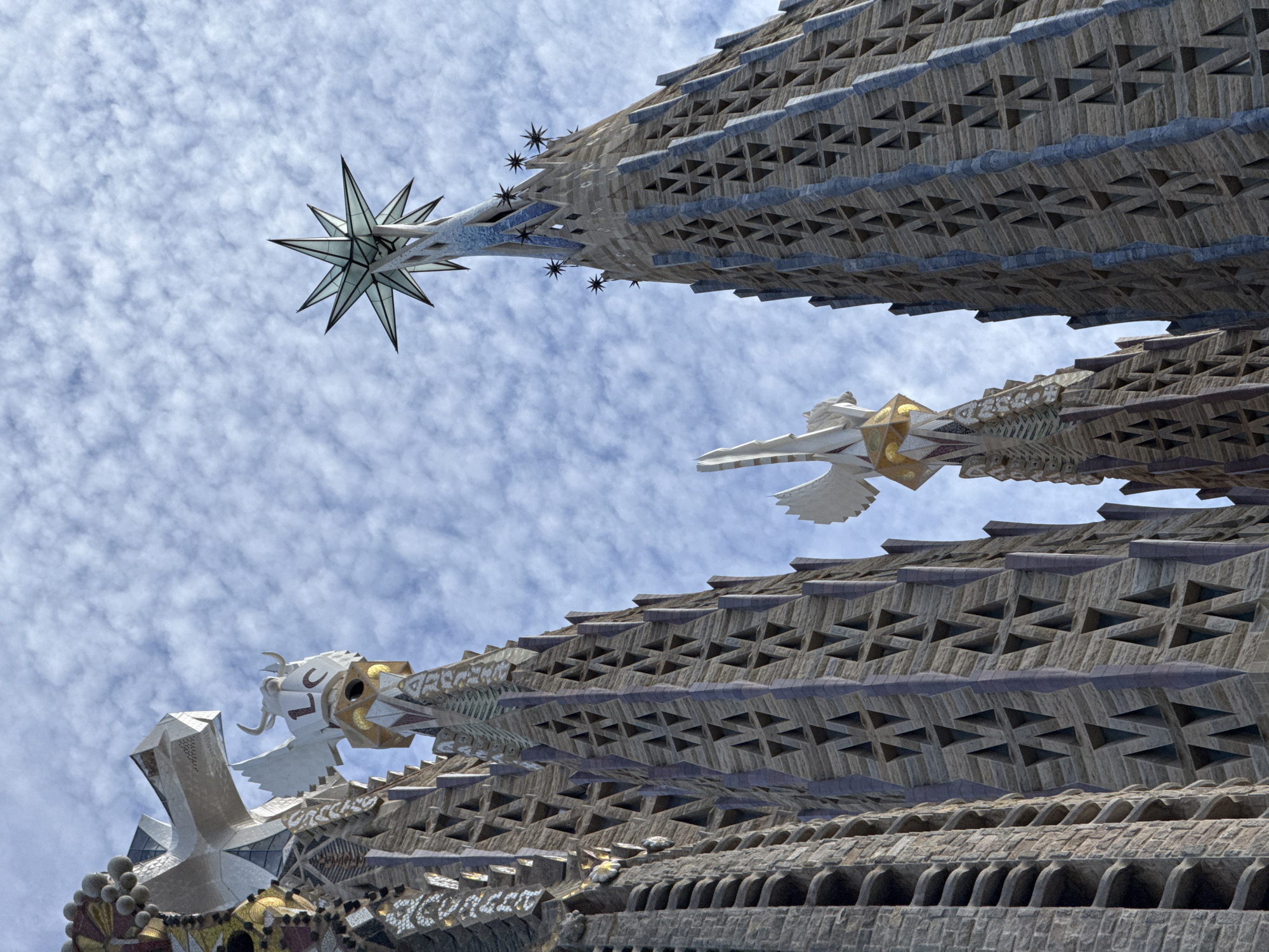 The image captures an upward view of the Sagrada Familia basilica in Barcelona, Spain, focusing on its intricate spires against a cloudy sky. The architectural style is highly ornate and Gothic, characterized by stone carvings and geometric patterns. One spire is topped with a large, multi-pointed star made of glass and metal, with smaller star-like ornaments adorning the edges of other spires. Other spires feature sculptural elements, including winged figures and what appears to be a bull's head with horns, adorned with the letters "L" and "C". The stone facades are covered in repeating triangular openings and textured surfaces, with accents of blue and gold. The lighting suggests it is daytime, with clouds scattered across a pale blue sky. There are no people visible in the image.