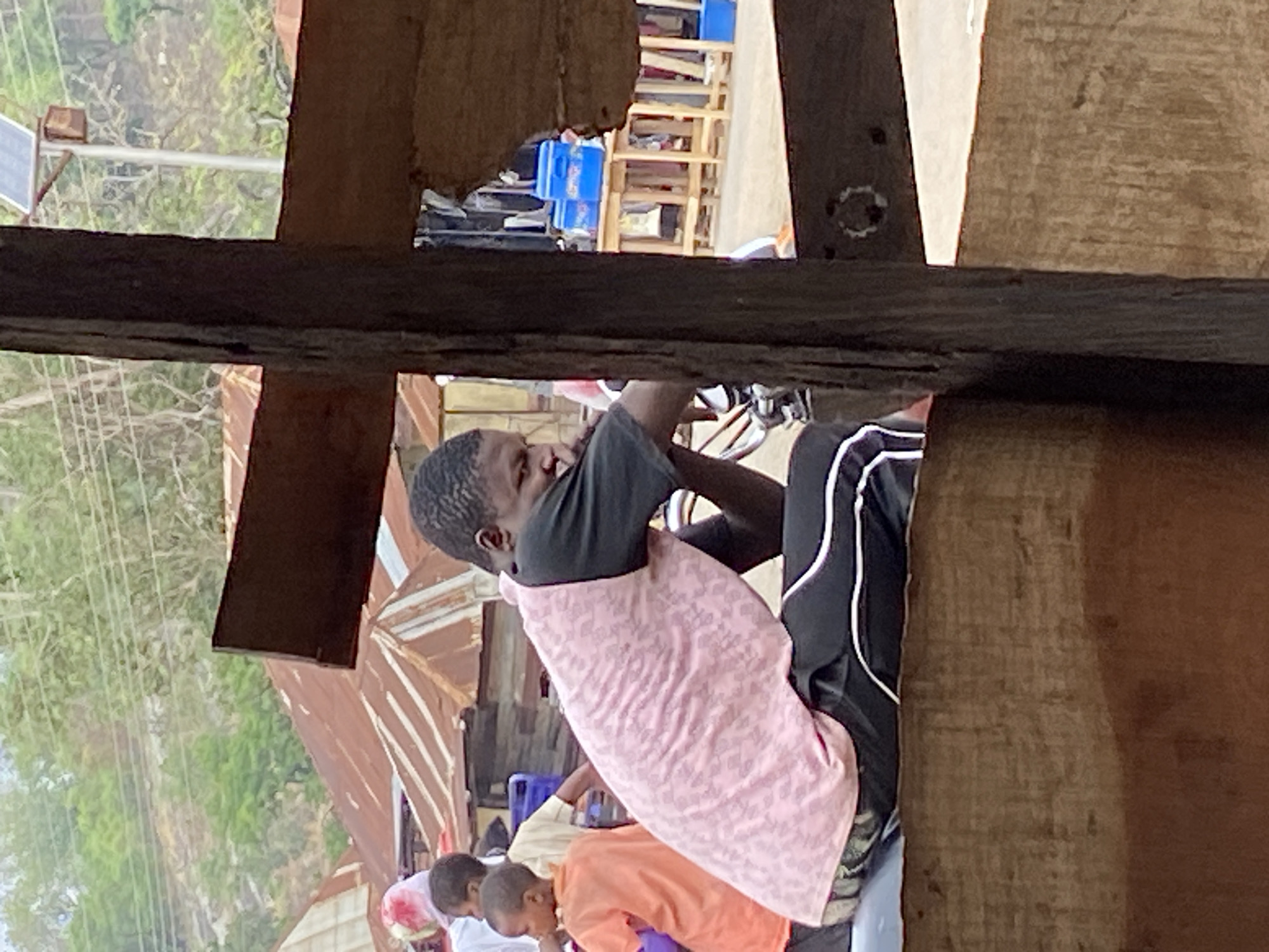 The image, taken in what appears to be Mararaba, Nigeria, captures a busy outdoor scene, framed by rough-hewn wooden beams that suggest a vantage point from within a structure or through a gap. The perspective is looking out towards a street or informal market area.
The primary subject is a young, dark-skinned man, seen from the waist up, wearing a black t-shirt and black trousers with white stripes. A pink patterned cloth is draped over his lap. He is leaning forward with his hands engaged, possibly working on or attending to something out of frame. Behind him, parts of a motorcycle, including handlebars, are visible.
Below the main subject, two other individuals are partially visible, both appearing to be younger, possibly children. One wears an orange shirt and the other a lighter-colored shirt, and both are bent over, seemingly focused on an activity on the ground.
The background is a vibrant mix of everyday street elements. To the man's left, a small solar panel mounted on a pole is visible against a backdrop of green trees and power lines. Further back, on the right, blue plastic containers are stacked on simple wooden shelving units. Corrugated metal sheets, likely forming roofs, are also visible, some appearing rusty. The overall lighting indicates clear daylight conditions. The scene suggests a semi-urban or rural environment with ongoing daily activities, possibly a repair shop or a vendor stall.