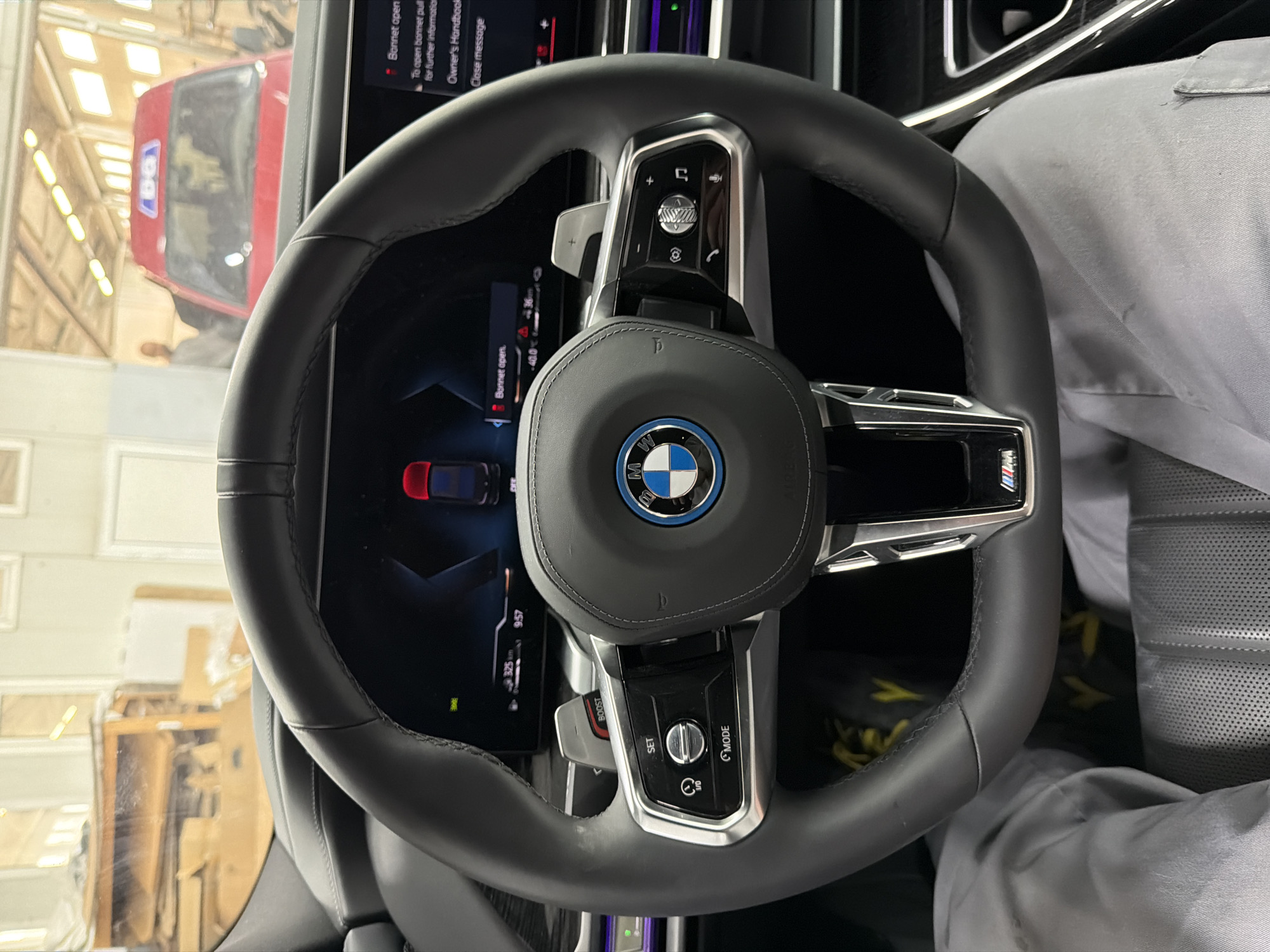 The image shows the interior of a BMW car, focusing on the steering wheel. The steering wheel is black leather with silver accents and features the BMW logo in the center. Buttons and controls are visible on the steering wheel's spokes. Behind the steering wheel is a digital display showing car information and a warning message about the bonnet being open. Through the windshield, a red van is visible, reflecting a warehouse-like setting with bright fluorescent lights and stacks of cardboard. The driver, wearing a gray shirt, is partially visible on the right side of the frame. The overall environment appears to be an automotive garage or service area.