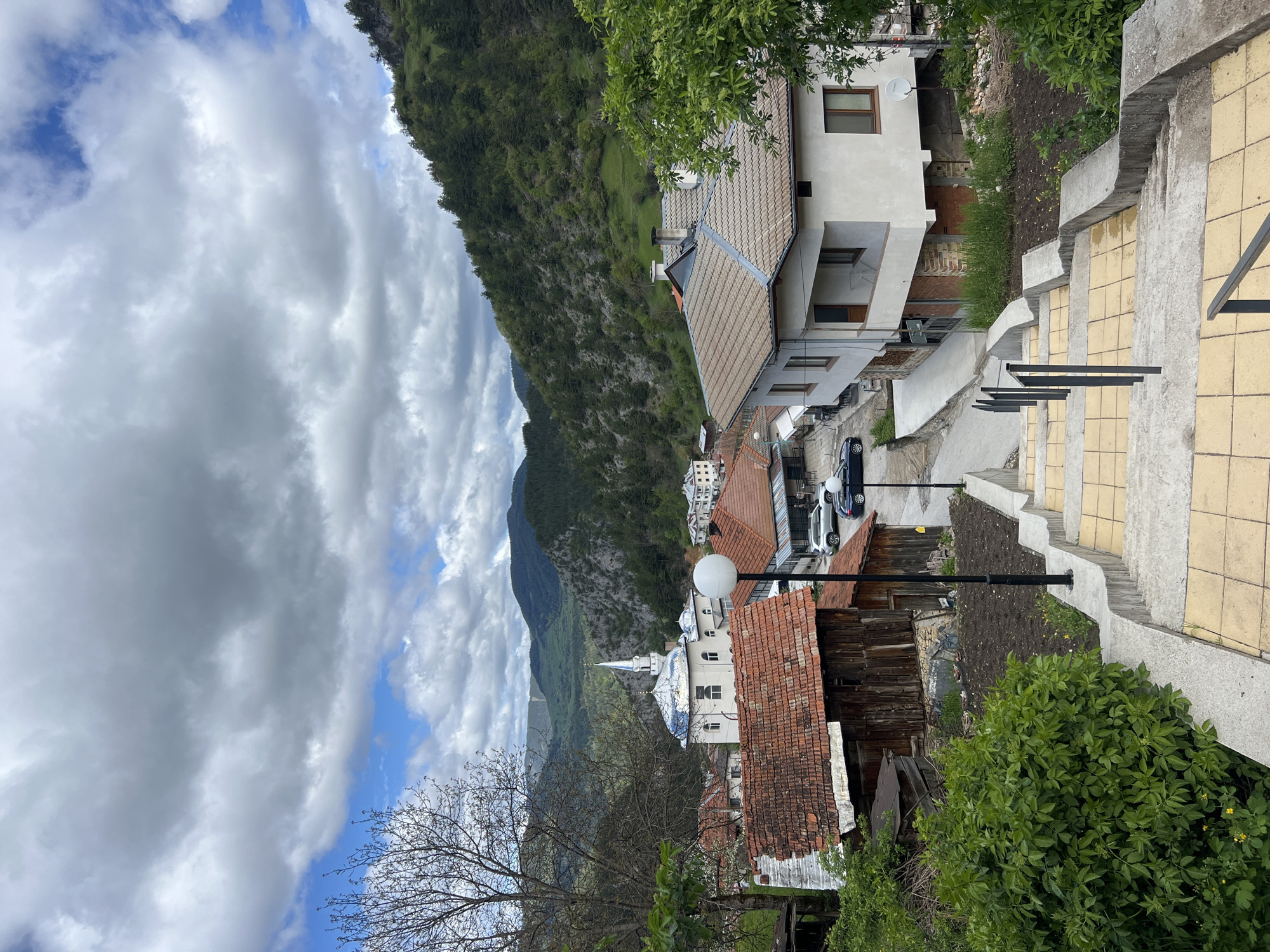 The image shows a view of the city of Giovren, Bulgaria, with a mountain range in the background and a cloudy sky overhead. The foreground features a set of tiled steps with black metal railings. To the left of the steps are various buildings with tiled roofs, including a white building with a spire. There is a mix of traditional and modern architecture. Bare trees and green foliage are interspersed among the buildings and along the edge of the frame. Cars are visible parked on a road between the buildings. Streetlights are also present.