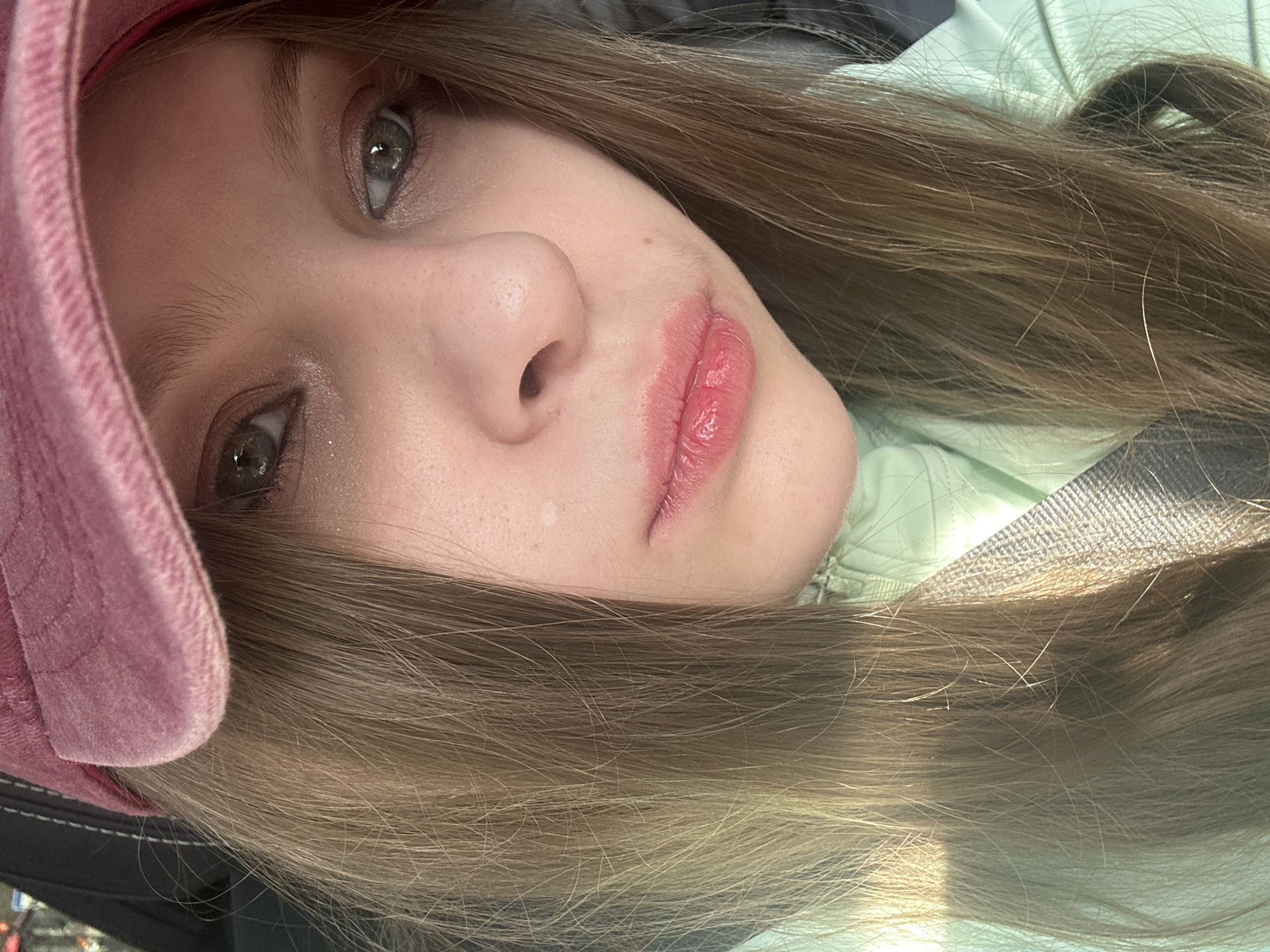 A close-up, tilted selfie of a young woman with light brown hair, wearing a pink hat and a light green jacket. She has fair skin, light-colored eyes, and pink lips. A safety belt is visible crossing her body. The background is blurred and does not provide enough detail to identify a specific location in Moscow, Russia. No specific actions or interactions are depicted.