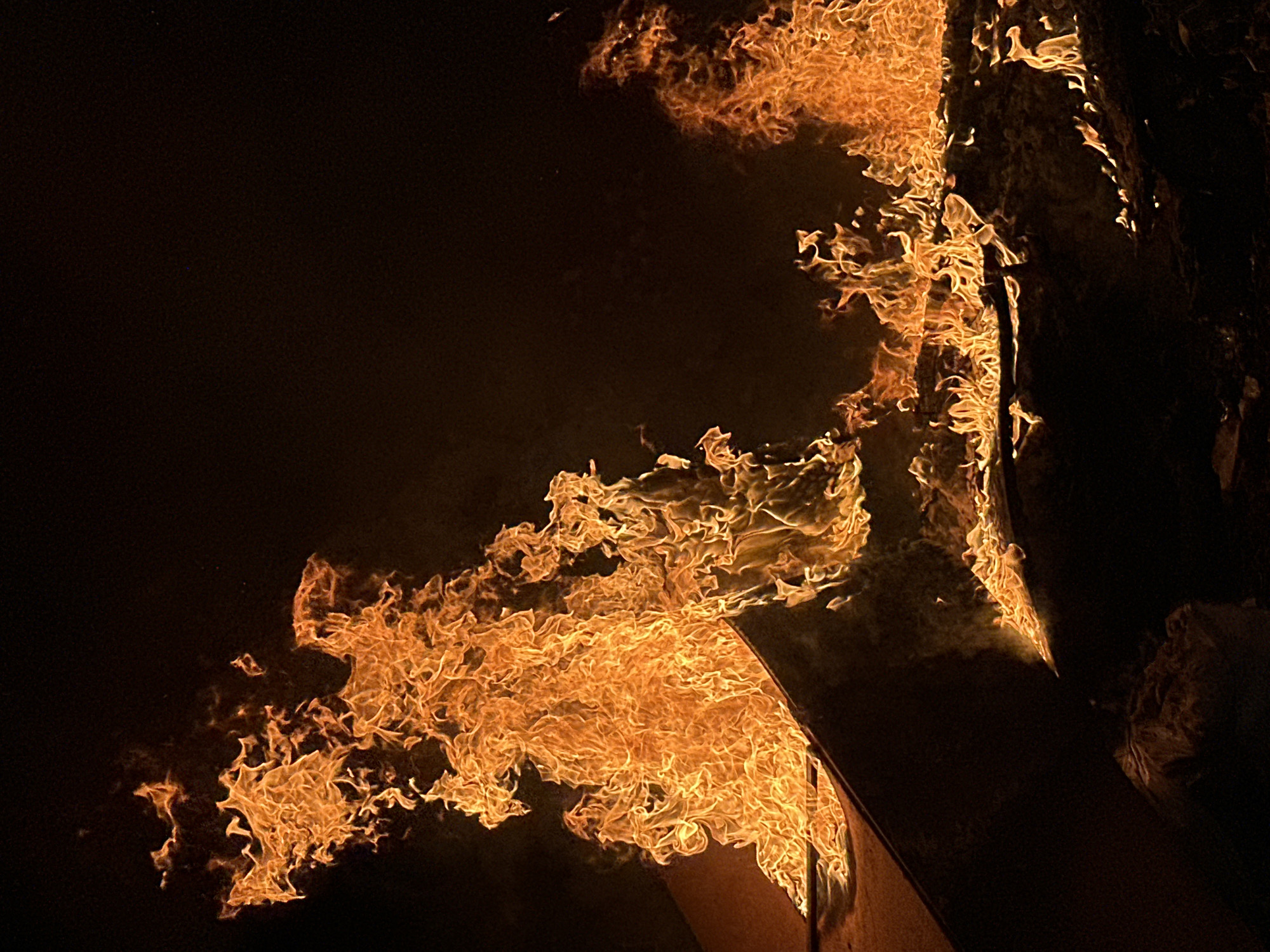 The image depicts a large fire occurring at night. The scene is dominated by bright orange flames licking upwards and to the left, consuming an unidentified dark object. The background is predominantly black, indicating darkness. Location and subjects are unknown.