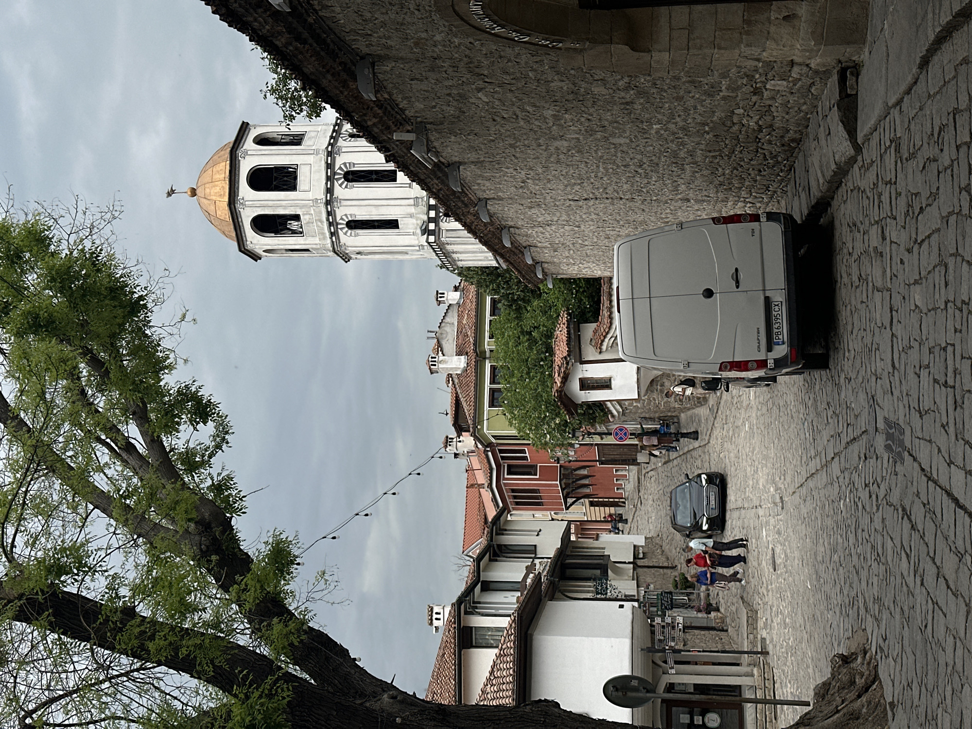 The image depicts a street scene in Plovdiv, Bulgaria, characterized by cobblestone paving and historic architecture. A white van is parked on the right side of the narrow street. The bell tower of the Church of St. Constantine and Helena is visible in the background, with its distinctive white walls and golden dome. Further down the street, a dark-colored car is parked and several people are walking and interacting. Buildings with tiled roofs and light-colored facades line the street. A tree with green foliage is in the foreground on the left. Overhead, lights are strung across the street. The sky is overcast.