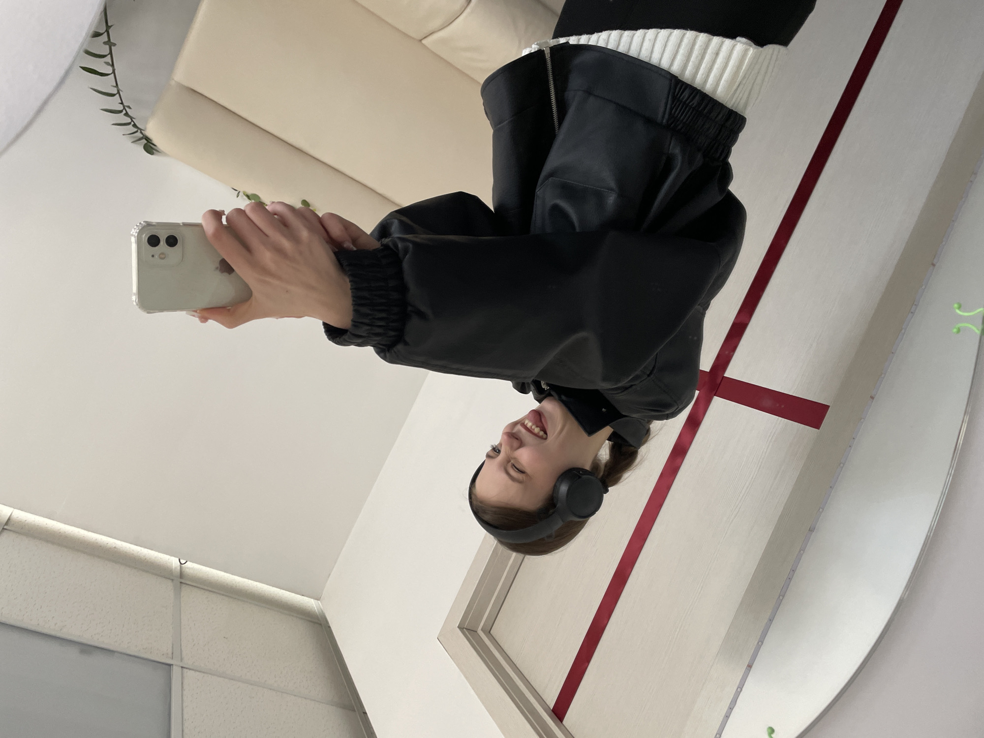 A female is positioned taking a selfie in front of a large mirror. She wears a black, leather jacket over a white ribbed sweater and black headphones. She holds a white smartphone in her hands. In the mirror's reflection is a beige couch, a white ceiling, and a wall with a red design. The photograph was taken in an indoor location and is likely not taken in the city of Фоки, Russia.