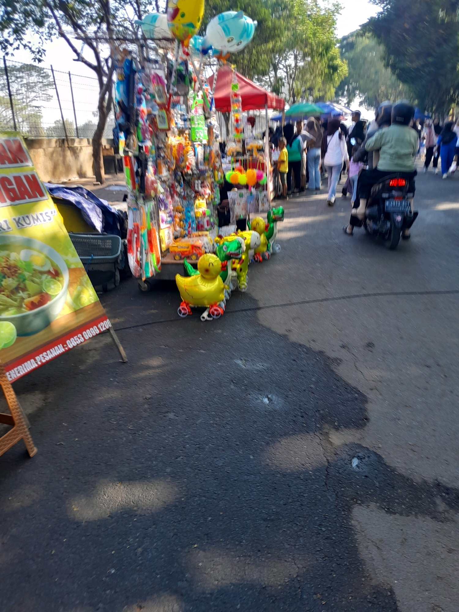 The image depicts a street scene in Kedungpedaringan, Indonesia, with a focus on a vendor's stall. The stall is laden with various items, including balloons shaped like ducks and dinosaurs, and other assorted toys and trinkets. In the foreground, a bright yellow duck balloon with wheels is prominently displayed. Behind the stall, people are walking along the street, and a motorcycle with two riders is passing by. The environment suggests an outdoor market or a busy pedestrian area, with trees lining the background and a sunny sky. A large yellow banner advertising food, possibly "Nasi Kunis," is partially visible on the left side of the image. The asphalt road surface is visible, with shadows cast by the sun and trees.
