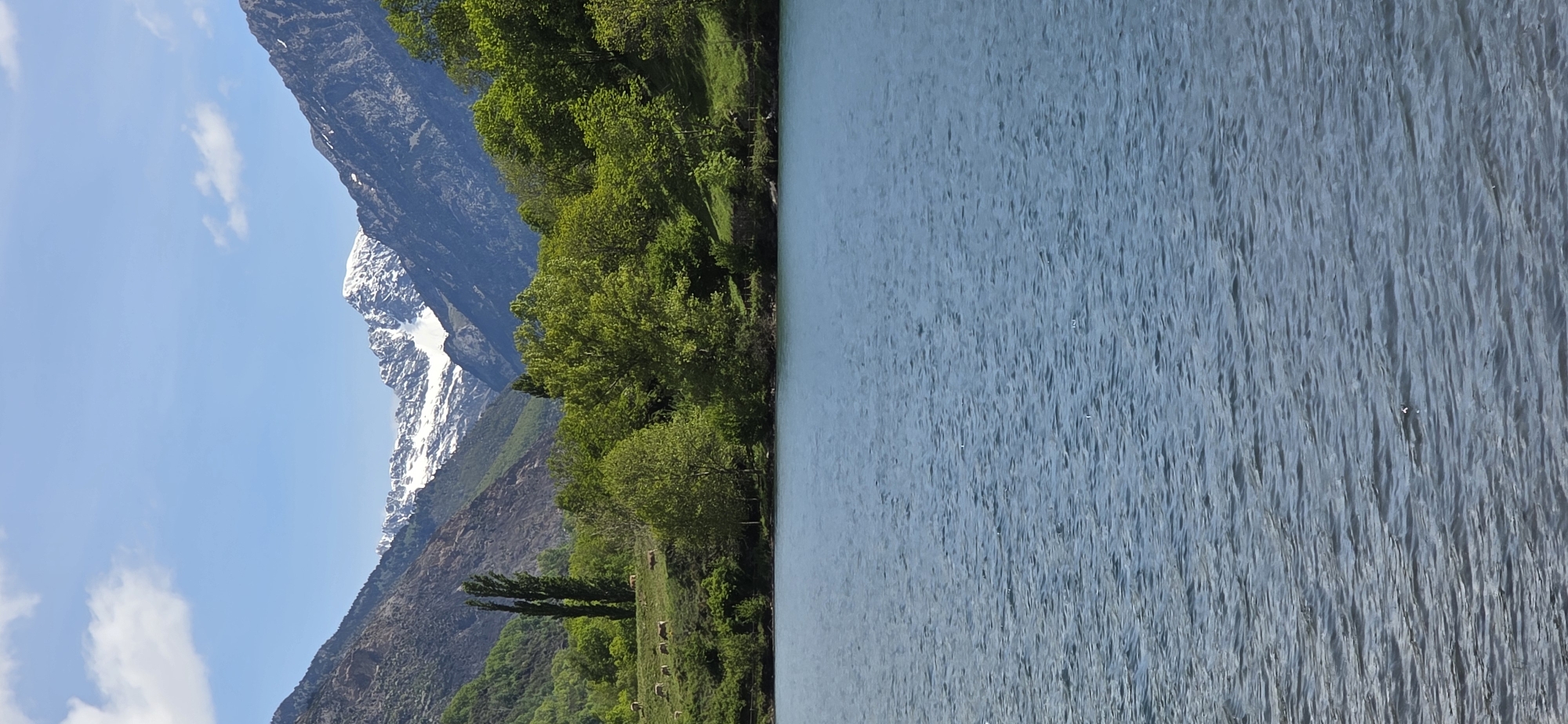 The image depicts a landscape in Sahún, Spain, showcasing a body of water on the right side and a mountain range to the left. A band of lush green vegetation, including trees, separates the water from the mountain base. The mountain's peak is partially covered with snow. The sky above the mountains is light blue with scattered white clouds. The water surface has a rippled texture. At the base of the mountain, there are several hay rolls.