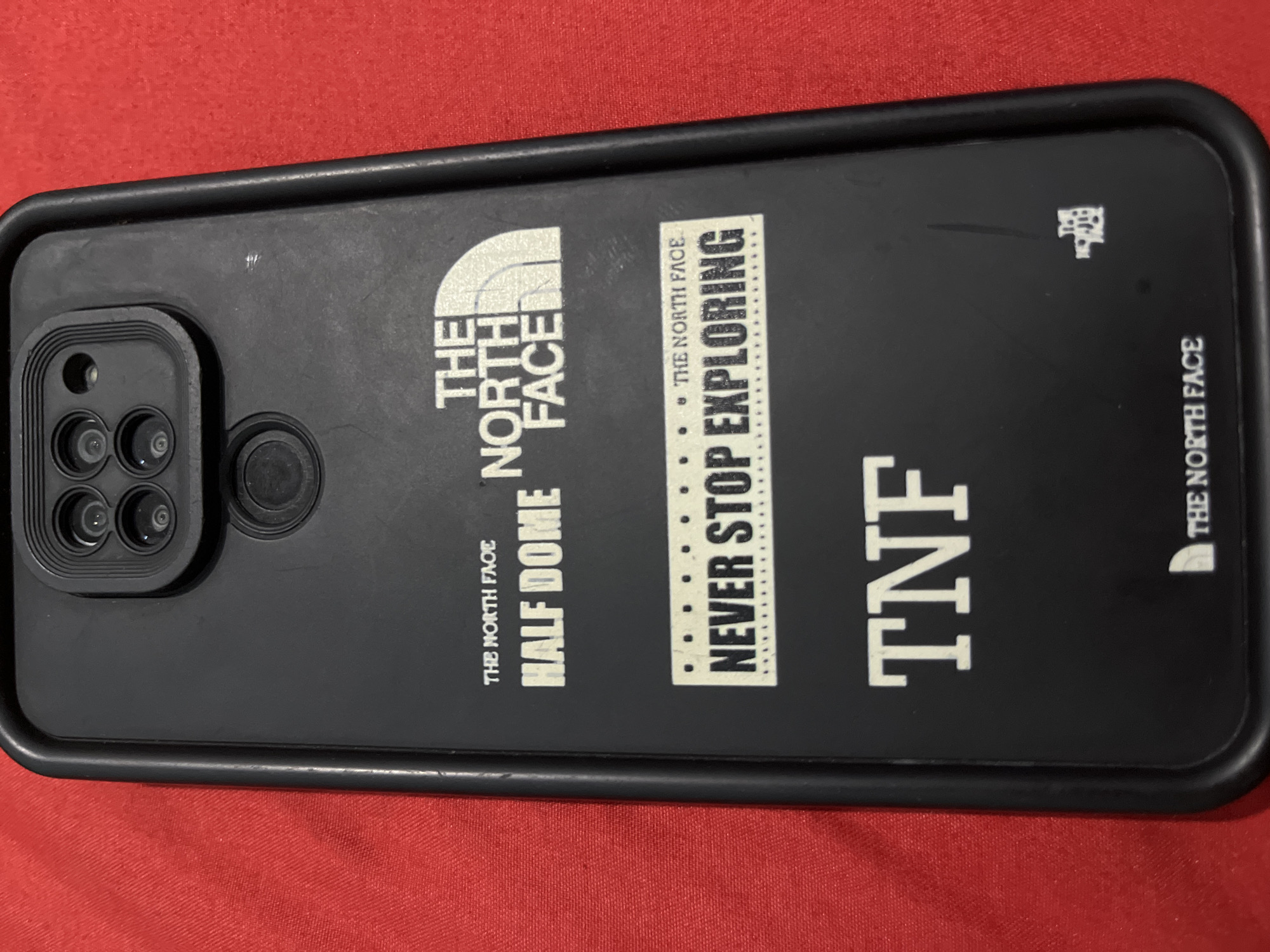 The image shows the back of a black phone case with the "The North Face" logo and text printed on it. The case is lying on a red textured surface, possibly fabric. The phone case features several distinct designs:

*   **Top:** A square camera module with four circular lenses and a circular flash.
*   **Mid-upper section:** "THE NORTH FACE" logo in white, with "HALF DOME" printed below it.
*   **Middle section:** A dotted line with small circles, followed by "THE NORTH FACE" and "NEVER STOP EXPLORING" printed on a rectangular white sticker-like design.
*   **Lower section:** "TNF" printed in large white letters.
*   **Bottom right:** A small white "The North Face" logo and "THE NORTH FACE" text printed horizontally.

The image does not provide any information about the location in Sumbernadi, Indonesia.