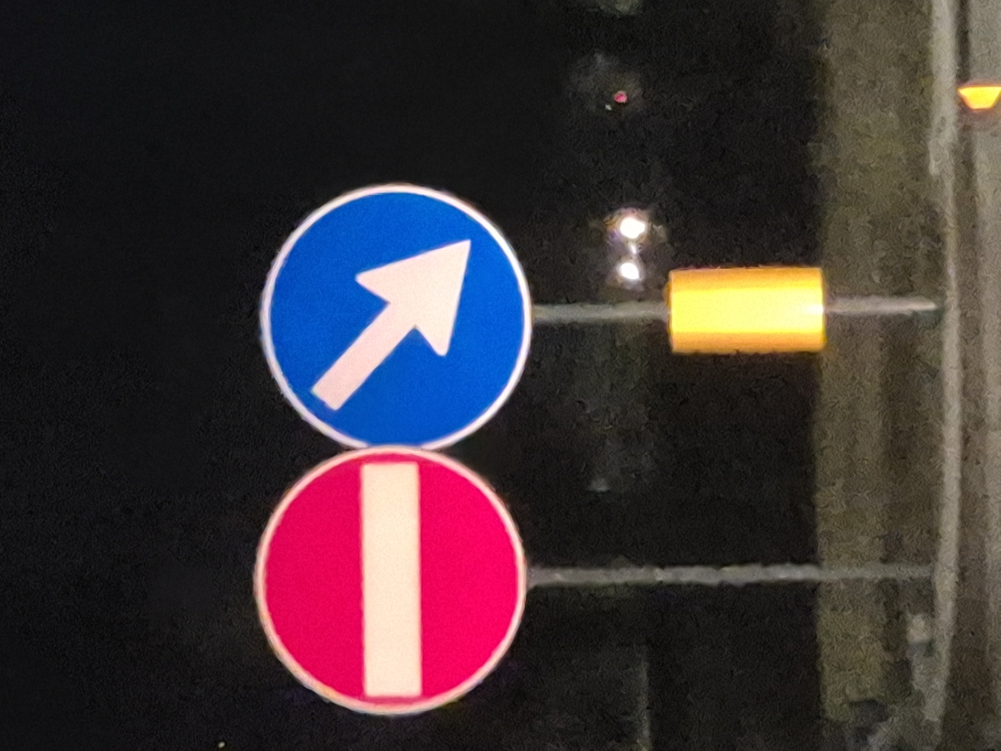 This is a nighttime, eye-level, medium shot of two road signs on poles in Taranto, Italy. The sign on the left is circular with a red border and a white horizontal bar, indicating "Do Not Enter" or "No Entry." The sign on the right is circular with a blue background and a white arrow pointing downwards and to the right, indicating "Mandatory Direction" or "Keep Right." Both signs are illuminated, likely by streetlights or vehicle headlights. A yellow, rectangular, illuminated marker is attached to the pole of the right-hand sign, just below the sign itself. The background is dark, suggesting it is night. The ground appears to be asphalt or pavement, with faint lines visible. There are some blurred lights and reflections in the background, possibly from other vehicles or distant buildings. No people are visible in the image.