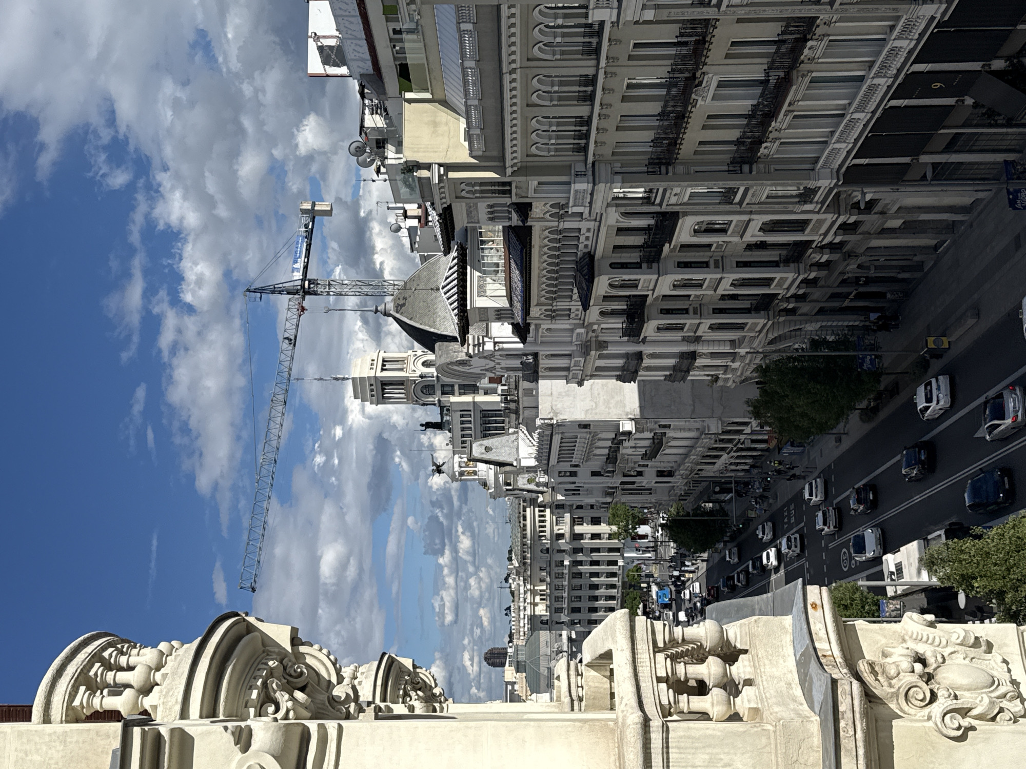 This photo shows a street in Madrid, Spain. The view appears to be from a high vantage point on a building, with ornate architectural details visible in the foreground. The street is lined with tall buildings, mostly light-colored with classical architectural styles. Traffic consisting of cars is visible on the street. A construction crane stands among the buildings, set against a backdrop of blue sky with white clouds. The scene appears to be during daylight hours.