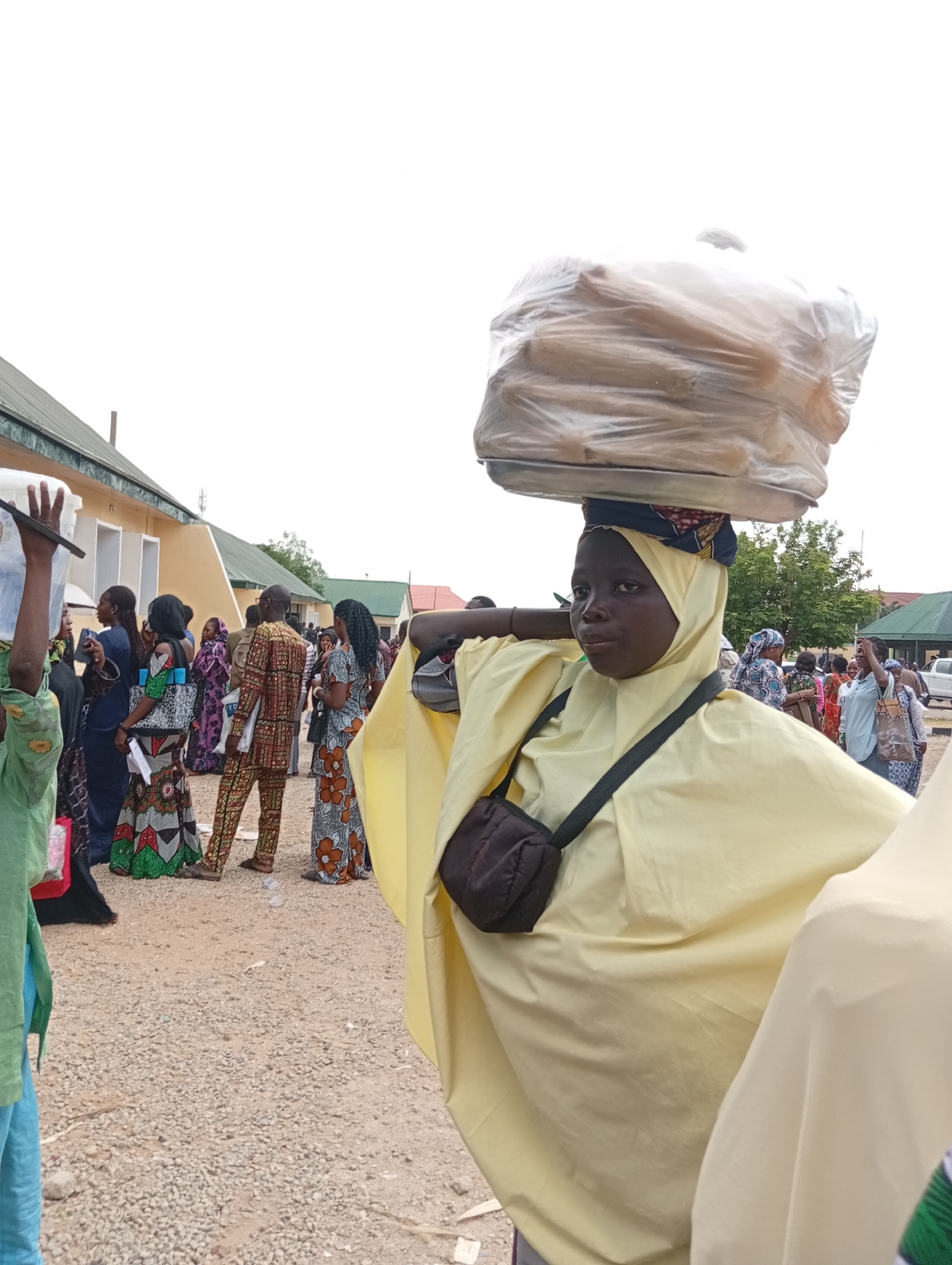 In Jalingo, Nigeria, a young girl, likely a vendor, carries a large load of bread wrapped in plastic on her head. She is wearing a light yellow headscarf and a matching flowing garment, with a dark cross-body bag. In the background, other people are visible, many in traditional African attire, suggesting a public gathering or market. Buildings with light yellow walls and green roofs line the area, and the ground is covered in gravel. The scene depicts everyday life in a Nigerian city.