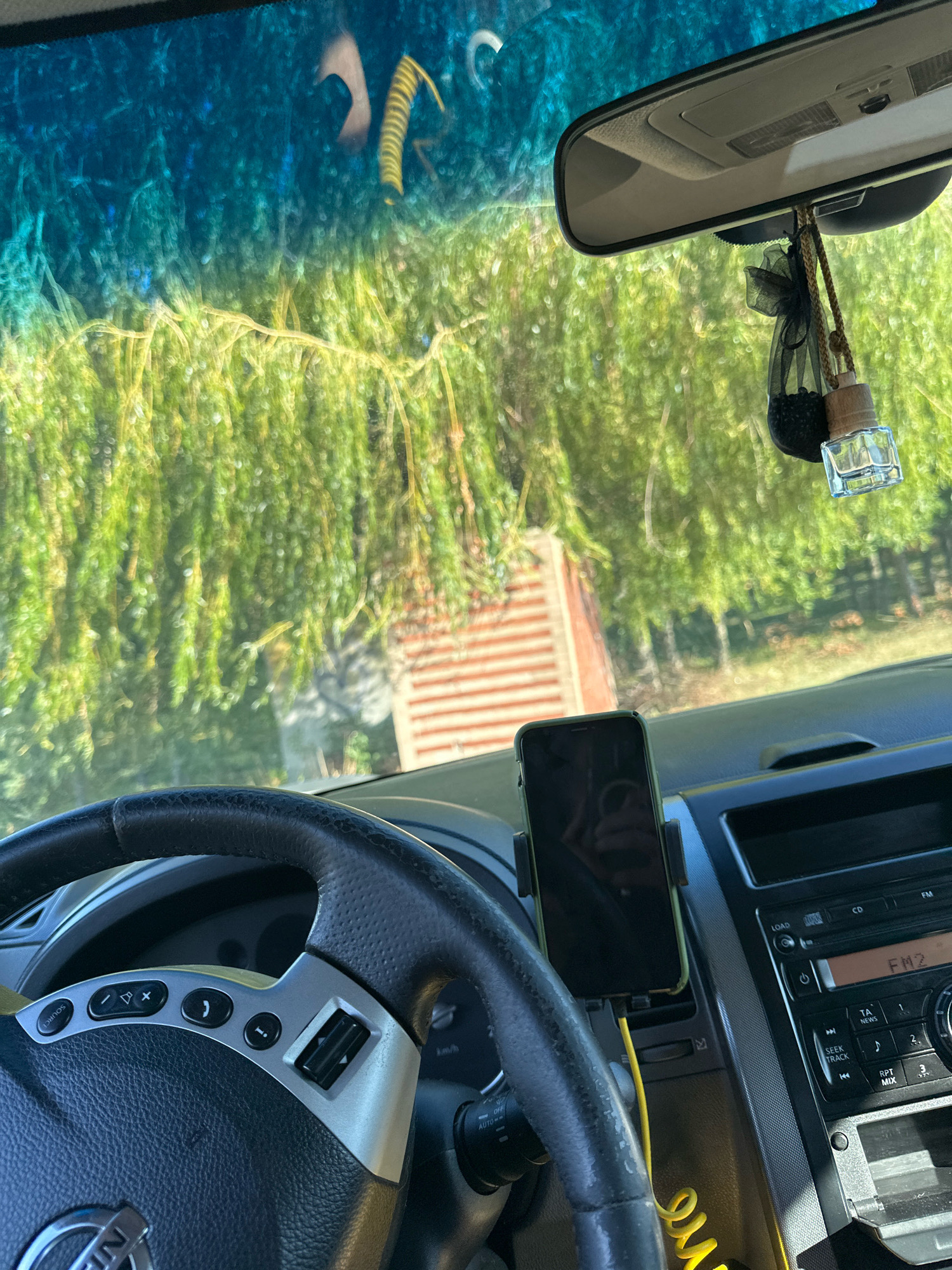 The image shows the interior of a car parked in Makiv, Ukraine. The view is from the driver's seat, looking forward through the windshield. Outside the car, a large willow tree with green leaves is visible, with a brick structure partially obscured by the foliage. The car's dashboard includes a steering wheel, a phone mounted on a holder, and a car stereo system. A car air freshener hangs from the rearview mirror. The sunlight casts shadows and highlights, suggesting it is daytime.