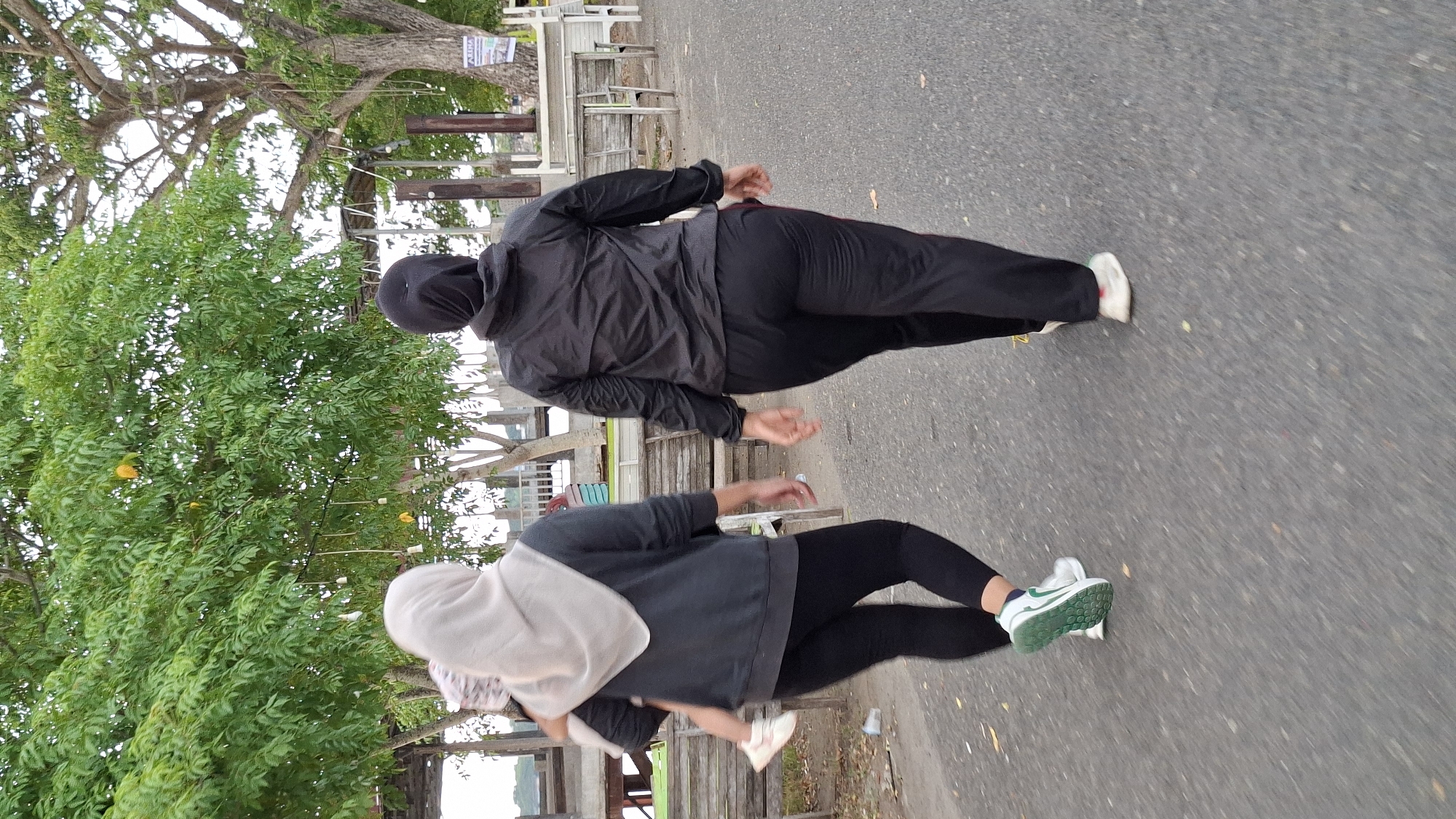 Two women in Islamic headscarves are walking away from the camera on a paved path, likely in Lhokseumawe, Indonesia. The woman on the left is wearing a light grey headscarf, a dark grey sweatshirt, and black leggings, and she is carrying a young child. The woman on the right is wearing a black headscarf and a black athletic jacket with black pants. They are walking in front of a lush green tree and a wooden structure with tables and chairs, suggesting they are in a park or a recreational area. The ground is a paved asphalt surface with some scattered leaves.