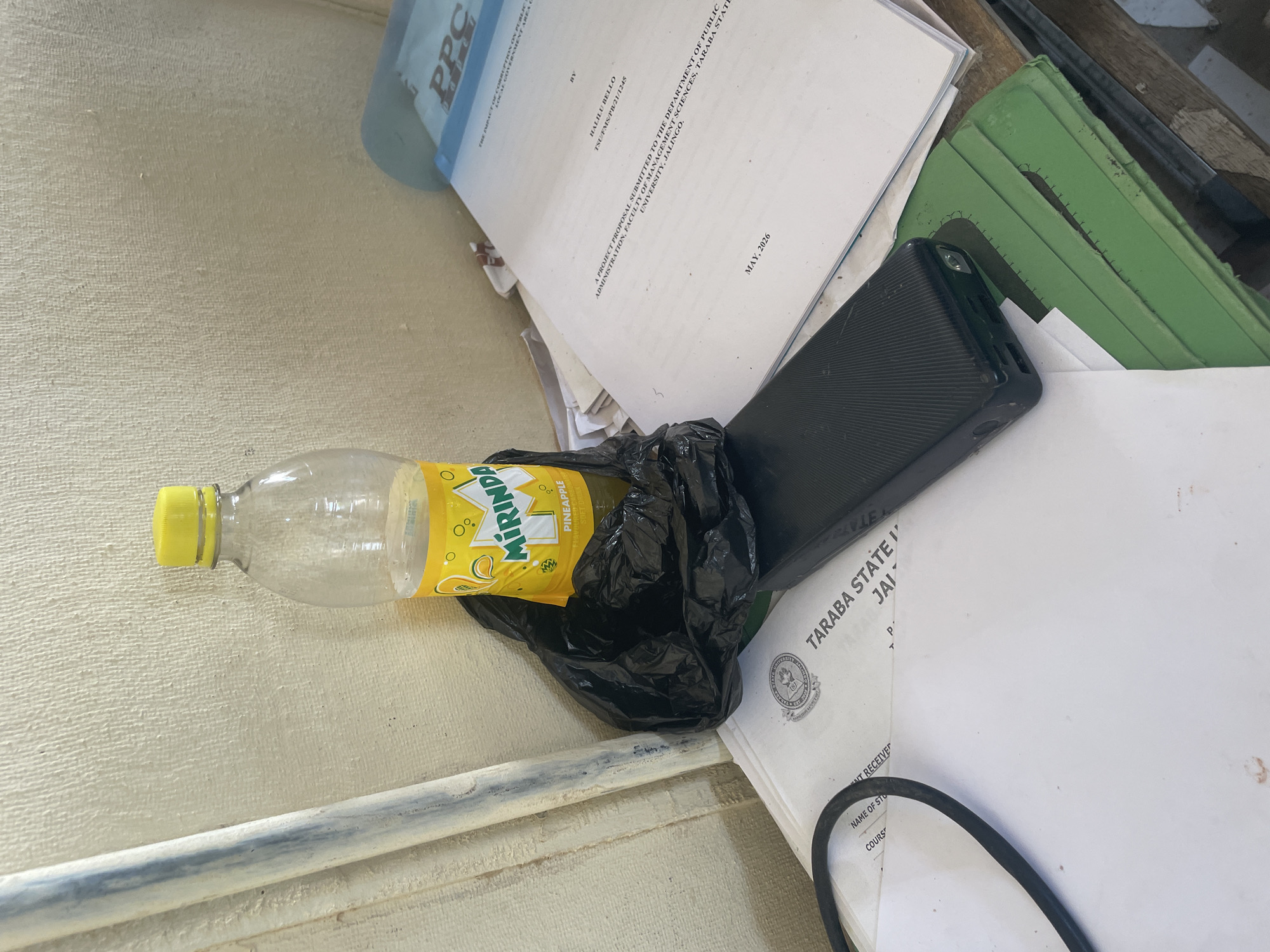 The image depicts a cluttered desk or surface with various items. The primary subjects are a partially consumed bottle of Mirinda pineapple-flavored soft drink, a black power bank, and several documents. One document is a project proposal with the title "A PROJECT PROPOSAL SUBMITTED TO THE DEPARTMENT OF PUBLIC ADMINISTRATION, FACULTY OF MANAGEMENT SCIENCES, TARABA STATE UNIVERSITY, JALINGO." This document is dated "MAY, 2026" and authored by "HALILU BELLO." Another document has "TARABA STATE" and "JAI" visible. A blue folder with "PPC" printed on it is also present. The scene appears to be indoors, possibly an office or study area, with a textured wall in the background and a metal pipe running along the bottom. The lighting suggests it could be daytime. There is no clear indication of an event taking place, but the arrangement suggests recent use or ongoing work. The overall impression is one of disorganization.