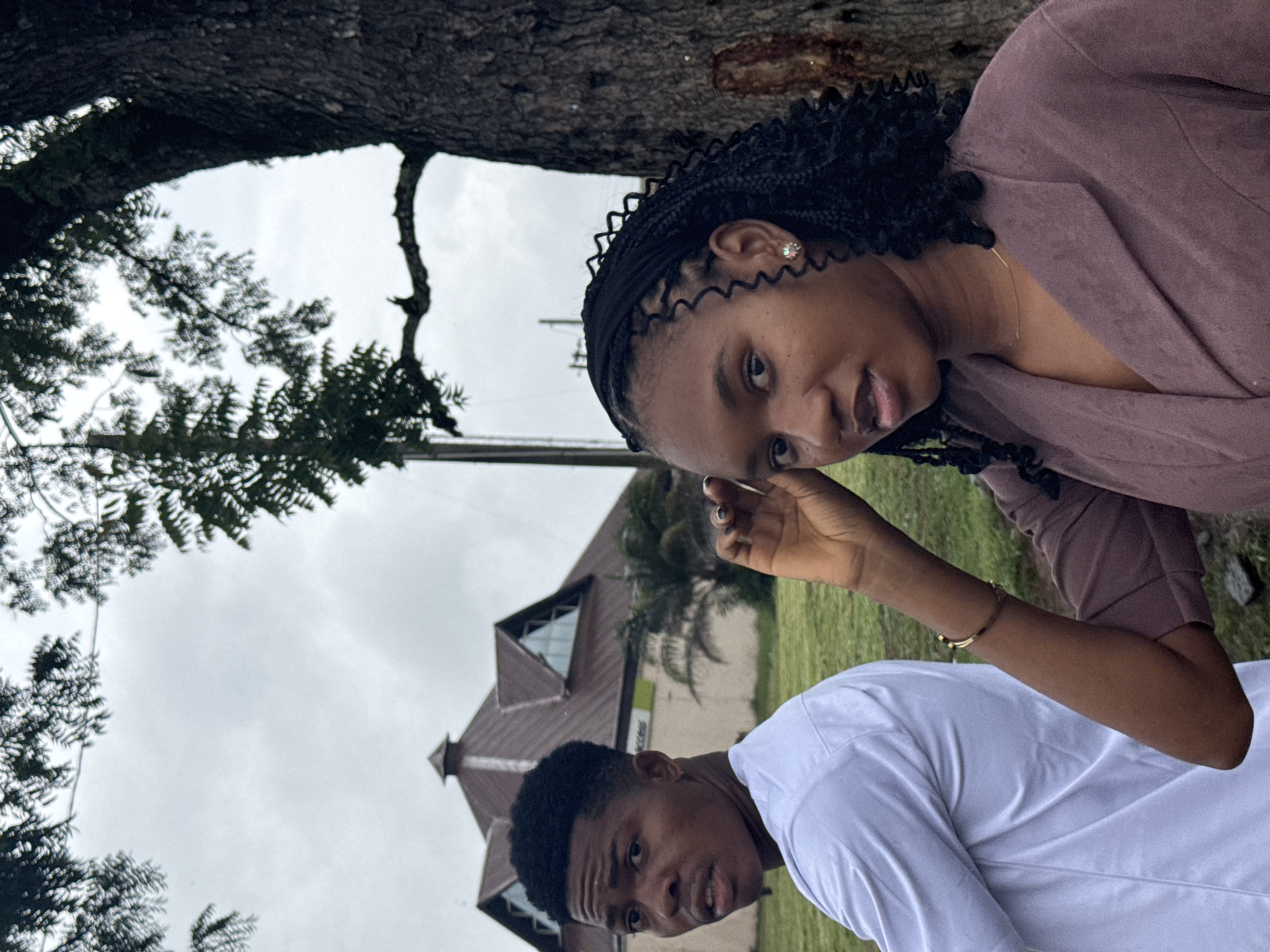 This image depicts two young adults, a male and a female, positioned outdoors in Ihiagwa, Nigeria, under an overcast sky.
The male, visible on the left side of the frame, is seen from behind but has turned his head to look over his left shoulder directly towards the camera, with his mouth slightly open. He wears a white long-sleeved shirt and has short, dark hair styled with a fade. A small hoop earring is present in his left ear.
The female, positioned closer to the camera on the right, has dark braided hair and wears a purplish-brown blazer. She looks directly at the camera, with her left hand raised towards her head. She is adorned with a gold-colored bracelet on her left wrist, a delicate gold-colored necklace, and a light-colored stud earring in her left ear.
In the mid-ground, a distinctive building with a multi-gabled, dark brown roof and several attic windows is visible on a grassy expanse. A green sign bearing the text "ACCESS" is discernible on its facade. A large tree with textured bark occupies the far right of the frame, partially obscuring the female, with its branches extending into the cloudy, light grey sky. Additional green vegetation, including a small palm tree, is present around the building.