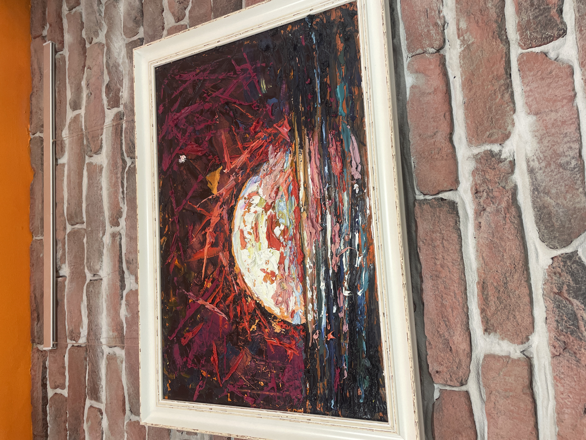 A framed painting of a sunset over water is hung on a brick wall. The painting features thick, impasto brushstrokes in vibrant reds, oranges, and yellows, with a prominent white and pale pink orb representing the sun partially submerged in dark, choppy water. The frame is a distressed white. The brick wall is made of reddish-brown bricks with white mortar. Above the brick wall, there is an orange ceiling. The scene is photographed from a slightly elevated angle, looking down at the painting. No people or animals are visible. The setting appears to be indoors, potentially a restaurant or gallery, given the decorative elements. The image does not provide any specific information about the city of Burgas, Bulgaria.