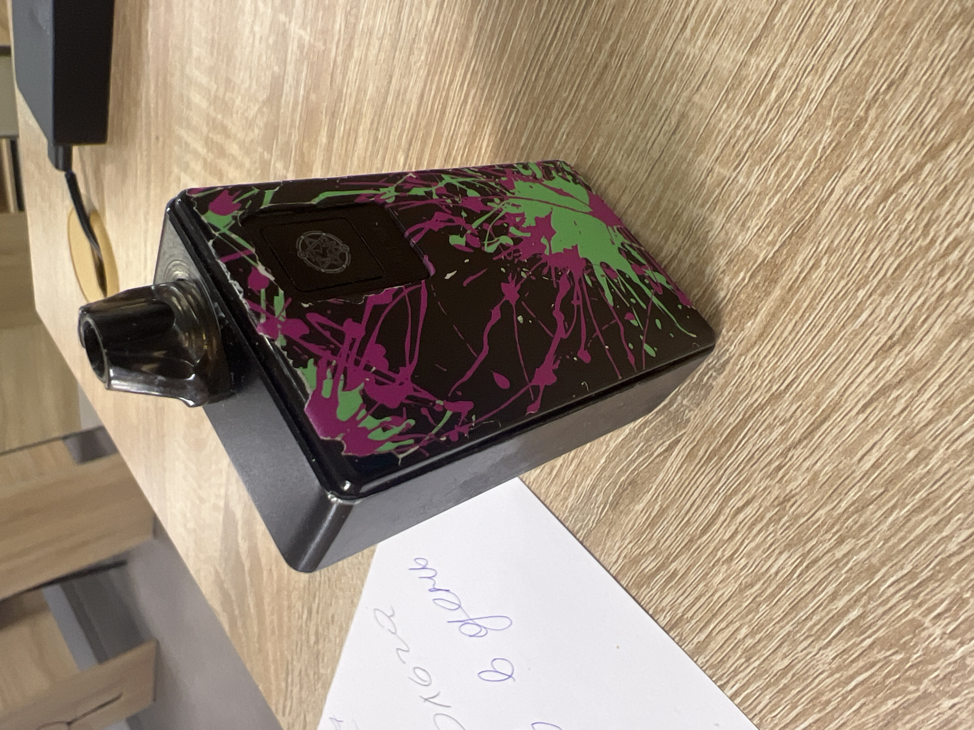 A black vape device with a splattered design of pink and green paint is positioned on a wooden table. A piece of white paper with handwritten Cyrillic text is partially visible to the left of the vape. In the background, out of focus, are wooden chair legs and a black charging brick connected to a cable. The image is a close-up shot, with the wooden grain of the table prominent in the foreground and midground. The scene is indoors, likely on a desk or table. There is no indication of the city of Minsk, Belarus within the image itself.