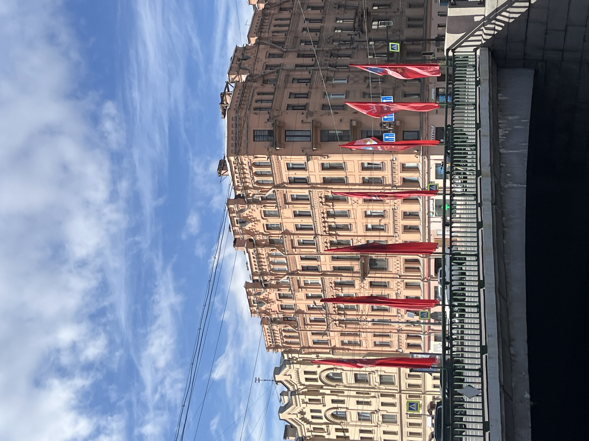 The image shows a street scene in an unidentified city. The focus is on a large, ornate building with multiple floors and windows. Red flags are suspended from a bridge or railing in front of the building. The sky is blue with scattered clouds. Power lines are visible against the sky. The bridge or railing appears to overlook a dark space, possibly a canal or another street.