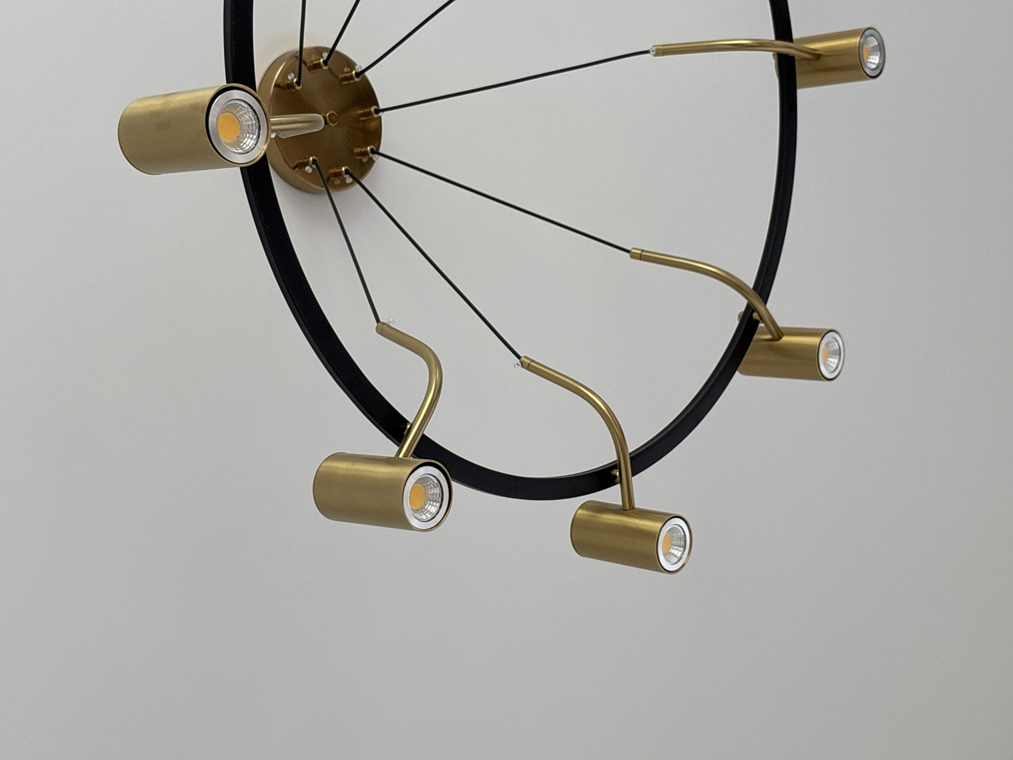 The image shows a modern, circular chandelier suspended from a gray ceiling. The chandelier has a black circular frame with gold-colored light fixtures attached to it at intervals. Each light fixture consists of a cylindrical housing with a visible LED light source. Thin black cables extend from the central, gold-colored mounting plate to the light fixtures, adding to the chandelier's unique design. The background is a plain gray ceiling, without any other visible details. There is no information to deduce the location as Ембаево, Russia.