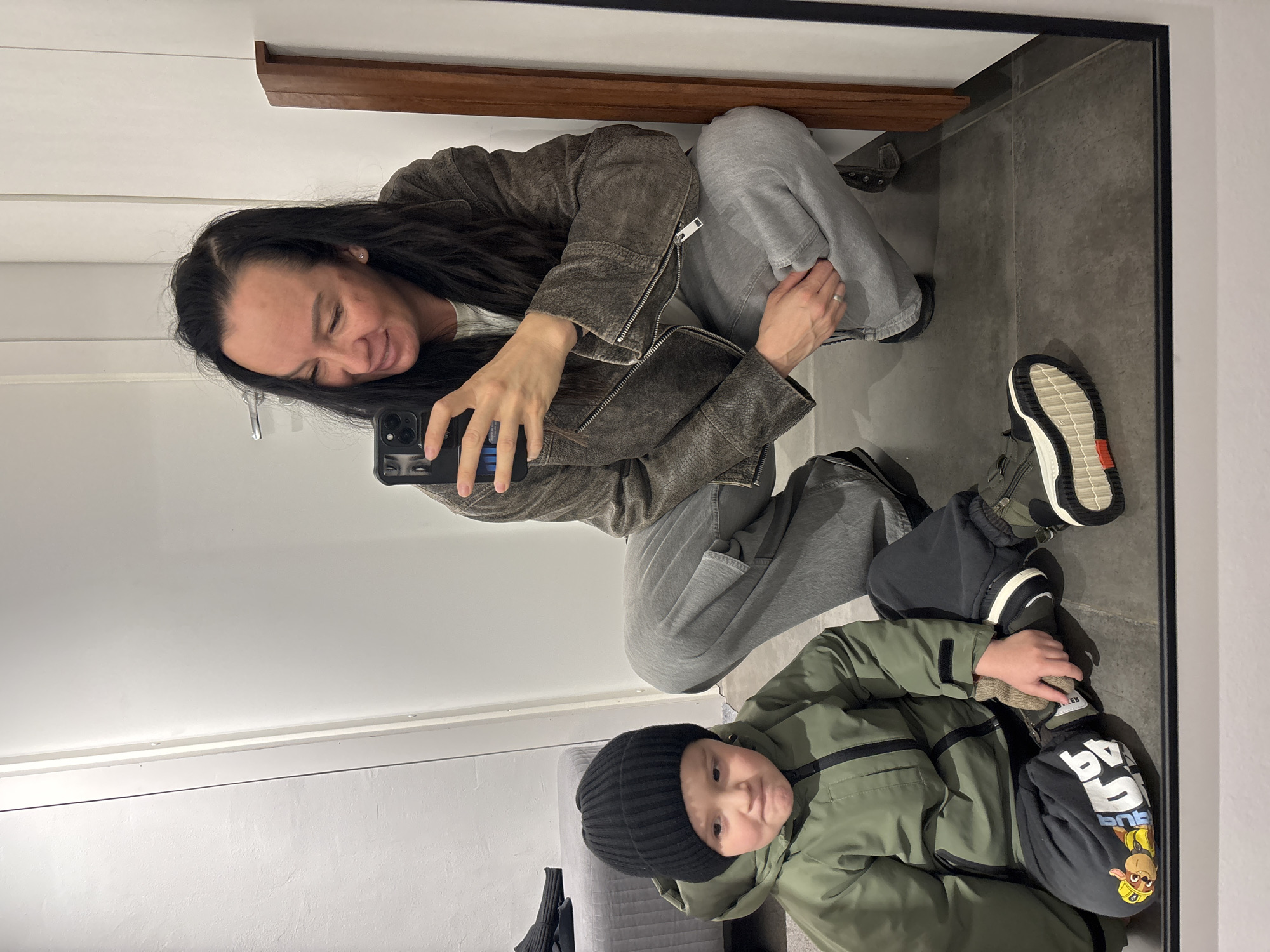 A woman and a young child are visible in a mirror reflection. The woman, wearing a leather jacket and gray sweatpants, is squatting and holding a mobile phone, presumably taking the picture. The child, wearing a black beanie and an olive green jacket, is lying on the floor. Their surroundings include a wooden shelf and a gray tiled floor. A Paw Patrol branded accessory is next to the child. The location is in Chaikovsky, Russia.