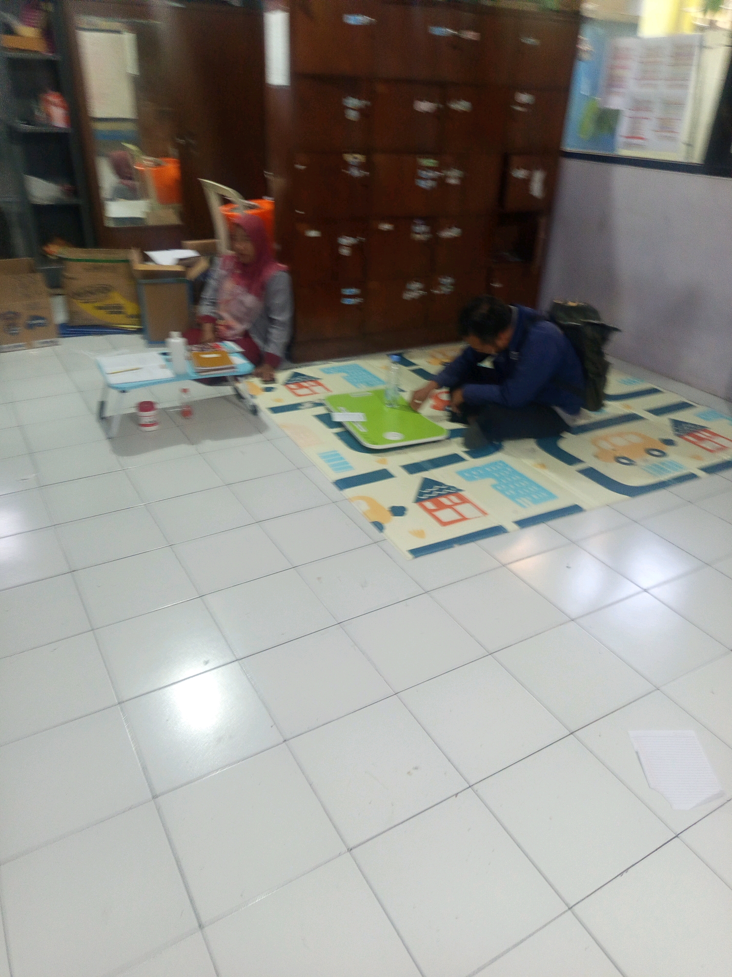 The image depicts an indoor scene with two individuals. On the left, a person wearing a pink headscarf and a grey shirt is sitting on the floor next to a small blue table, engaged with some items on the table. On the right, another person in a blue shirt is crouching on a patterned mat, possibly interacting with items on the mat or on a green tray in front of them. Behind the seated individuals, a tall wooden cabinet with multiple drawers is visible. The floor is tiled with white square tiles. The setting appears to be a room, potentially a classroom or a common area, and given the overall ambiance, it is likely located in Malang, Indonesia.