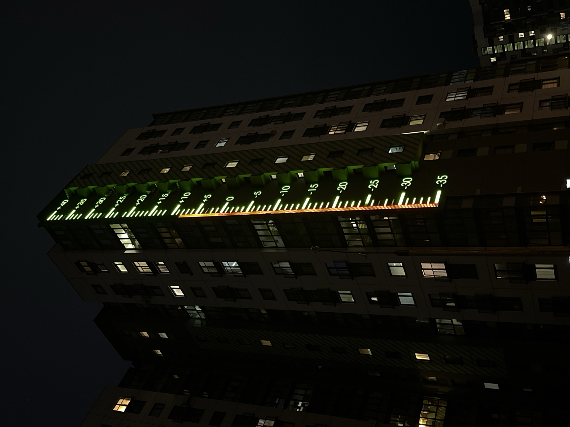 A low-angle shot captures a tall building at night, seemingly in a city, judging by the other building faintly visible in the upper right corner. The focus is on the front of the building, where a large, glowing thermometer is installed horizontally. The thermometer is marked with a scale ranging from +40 to -35 degrees, with each increment labeled. A red line indicates the temperature, falling somewhere between +5 and 0 degrees. The building itself is multi-storied with numerous windows, some lit up, indicating occupancy. The sky is dark, and the bottom half of the image appears to be a reflection of the building, possibly on a wet surface. The perspective is skewed, adding a dynamic angle to the composition.
