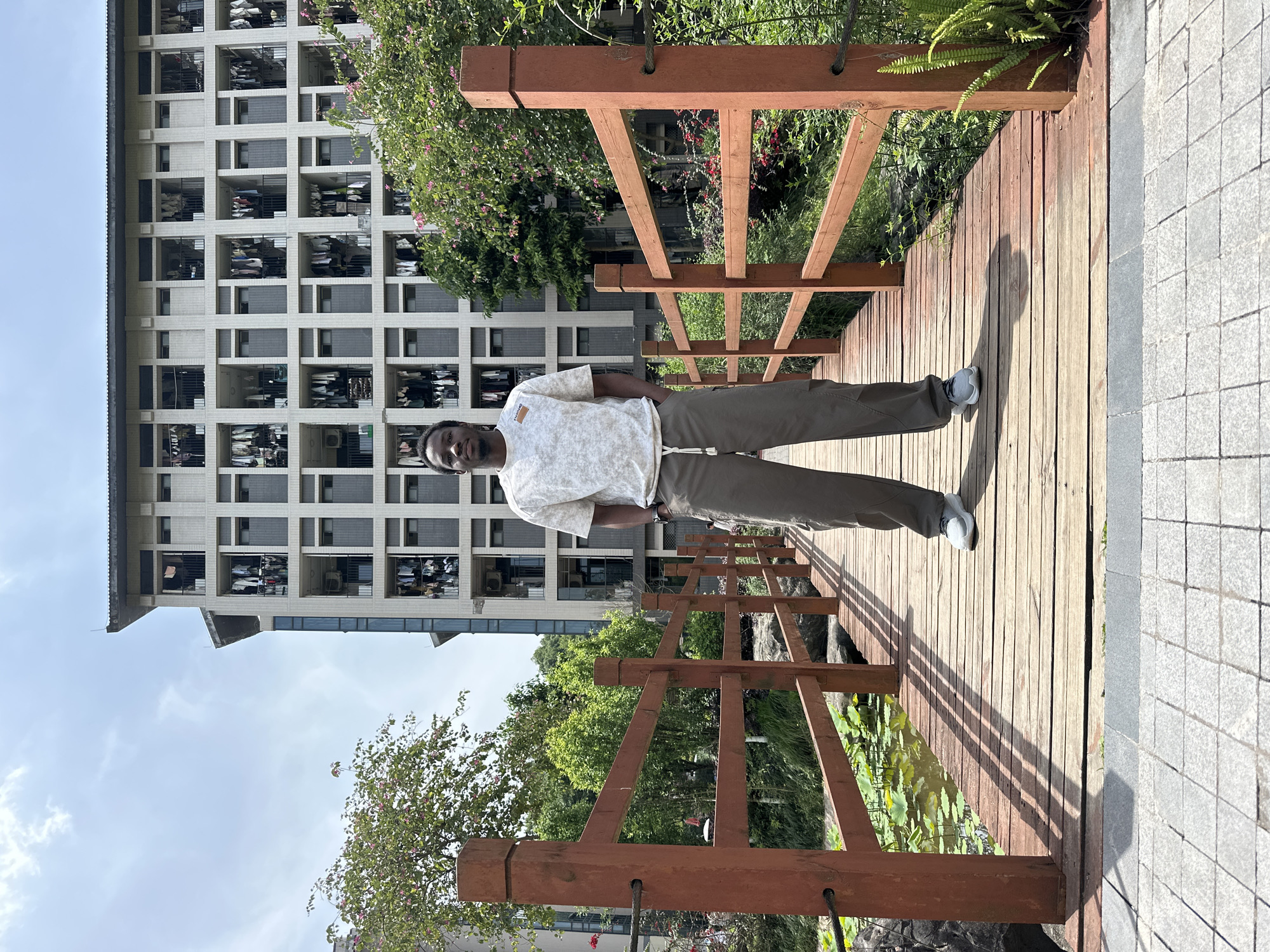 A man stands on a wooden bridge in Jiangjin, China. The bridge is constructed with wooden planks for the deck and dark wood railings on either side. Underneath the bridge, there is a view of greenery and water. In the background, a tall, multi-story apartment building with numerous windows and balconies is visible. The building is light in color and the balconies appear to have laundry hanging out to dry. The sky is partly cloudy. The man is wearing a white shirt and khaki pants, and he has his hands in his pockets. He is standing directly in the center of the bridge. The scene is bathed in natural daylight.