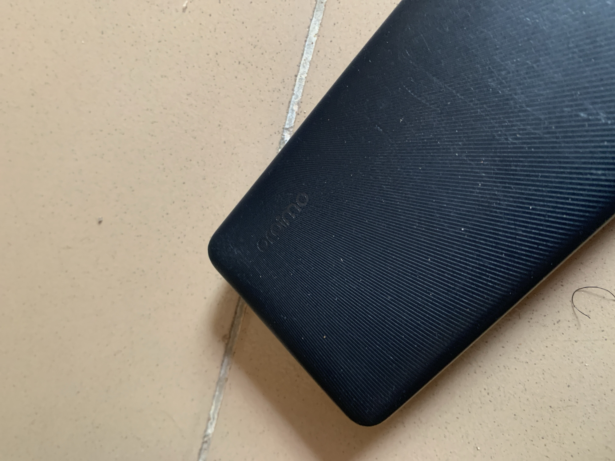 A dark blue, rectangular power bank with a ribbed texture rests on a light beige, tiled surface. The power bank is angled with its top left corner closest to the viewer. Faint, embossed lettering appears on the power bank, possibly a brand name, but it is not clearly legible. The tiled surface shows a thin, white grout line running diagonally across the image, separating two tiles. A shadow of the power bank is cast to its left. There are no people or any other objects visible in the immediate vicinity. The scene appears to be indoors, in Jauro Yinu, Nigeria.