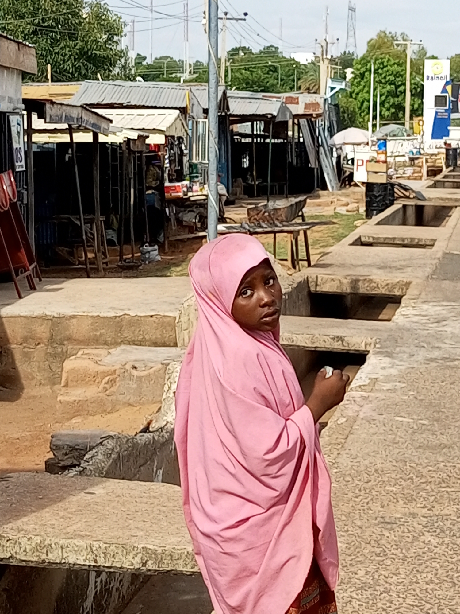 A young girl wearing a pink hijab and a patterned skirt stands in an outdoor market setting in Gombe, Nigeria. She is looking over her shoulder towards the camera with a slightly curious expression. Her right hand is raised and appears to be holding something small.
The background reveals a bustling market scene. Stalls with corrugated metal roofs are visible, some appearing to be selling various goods. Wooden structures and tables are set up, and in the middle ground, a wheelbarrow is resting on a table. A white and blue sign with the text "Rainoil" and a yellow circular logo is also visible in the background, suggesting a commercial area. The ground is a mixture of packed dirt and paved surfaces, with concrete structures and channels running through the scene.
The lighting suggests it is daytime, likely late morning or afternoon, with clear skies indicated by the bright illumination and shadows. The overall impression is a candid shot capturing a moment in a lively local marketplace.