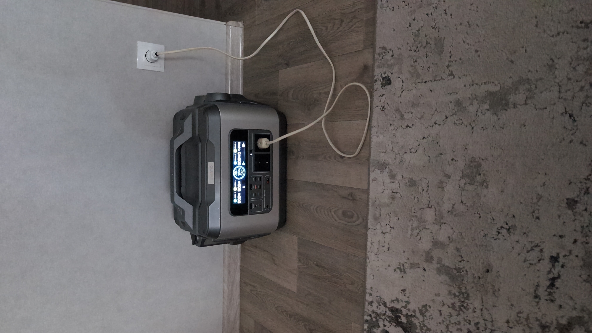 The image shows a portable power station plugged into a wall outlet on a wooden floor. The power station is silver and black with a digital display showing battery status and power output. A white cord connects the power station to the outlet. A grey and white rug is visible in the foreground. The scene is indoors, likely in a residential setting, with a plain grey wall in the background. There are no people or animals present. The image does not contain any information that would indicate its location in Kharkiv, Ukraine.