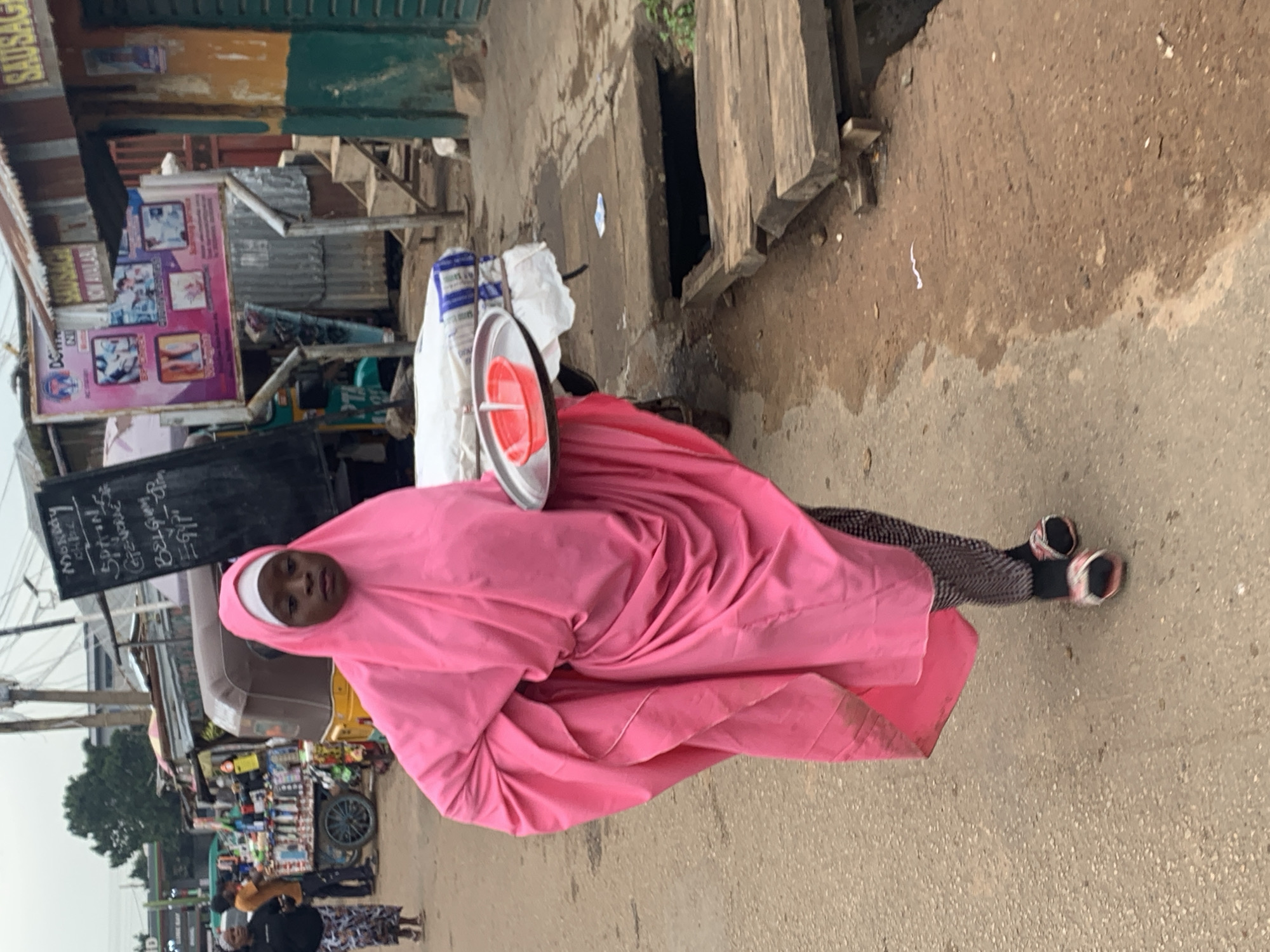 In the city of Jos, Nigeria, a young girl wearing a bright pink hijab and dress walks along a dirt road, carrying a tray of food. The girl, who appears to be of African descent, has her hair covered by the hijab, and her eyes are looking straight ahead. The dress is flowing and voluminous, and the girl is wearing a pair of patterned leggings and sandals. The tray she is carrying is silver, and it contains a red bowl with a white straw. On the tray, there are also some white packets that appear to be food items.
The girl is walking past a market stall that is selling various goods, including bags of rice and other food items. The market stall has a sign that says "Sausage Now Available." In the background, there are other market stalls and buildings, as well as a few people walking around. The road is unpaved and dusty, and there is a drain on the right side of the road. The overall impression is of a bustling market scene in a developing country.