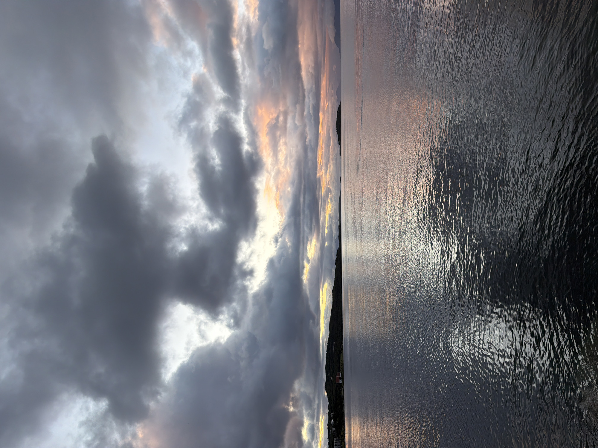The image depicts a serene, wide-angle view of the sea and sky at dusk, likely taken from the coast of Harstad, Norway. The sky is filled with dramatic, dark grey clouds, tinged with hues of pink and orange from the setting sun, which is partially visible through breaks in the clouds. These colors are reflected on the calm surface of the water, creating a shimmering effect.
In the middle distance, a tree-covered island or peninsula stretches across the horizon, with a few buildings, including what appears to be a red-roofed house, nestled along its edge. The water in the foreground is dark and reflects the sky's colors, with subtle ripples breaking the surface. The overall atmosphere is one of peaceful solitude and the natural beauty of a coastal landscape at the end of the day.