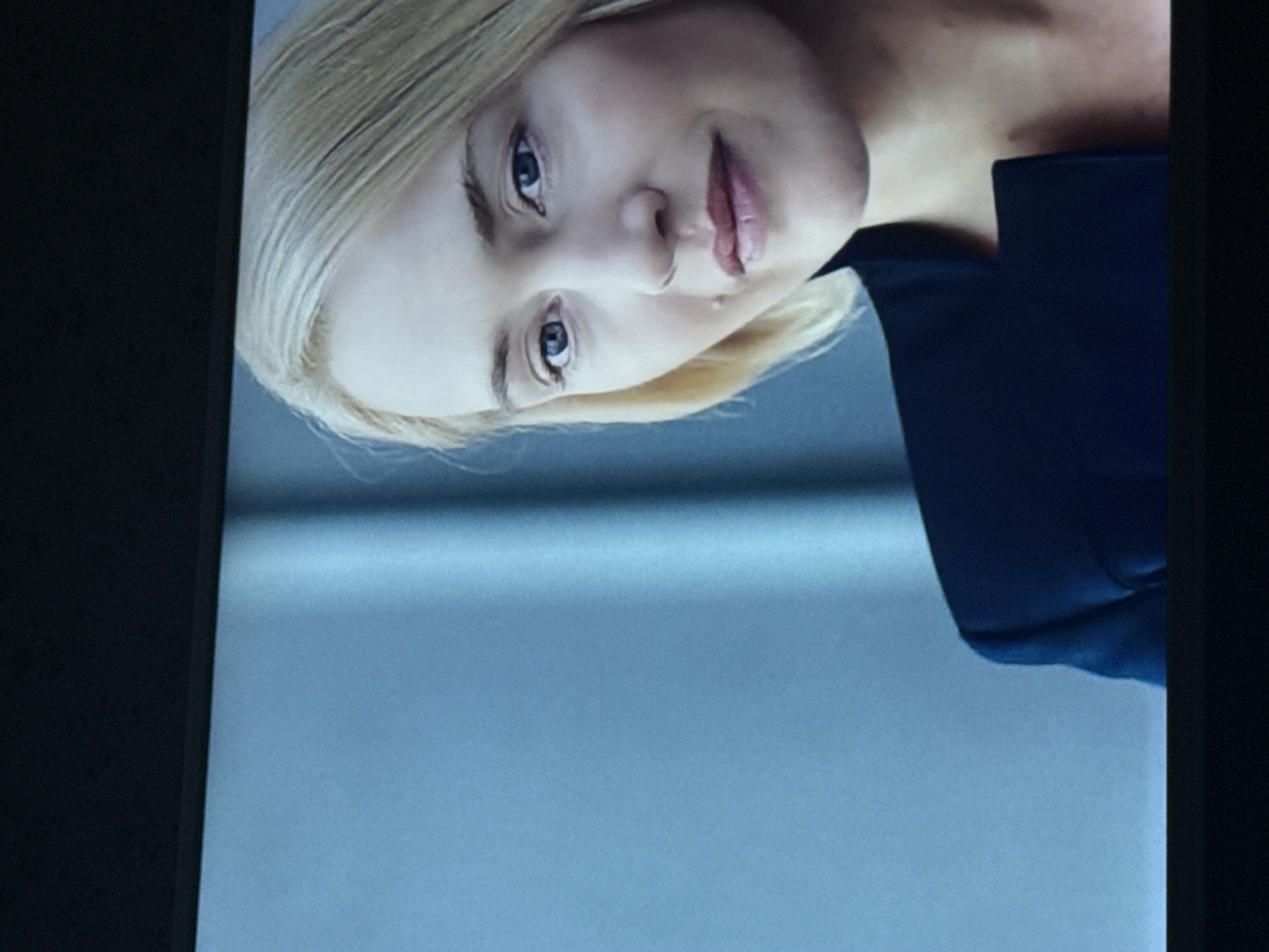 The image shows a close-up of a fair-skinned woman with blonde hair, blue eyes, and light pink lips. She is wearing a dark blue collared shirt. The background is a light blue-gray gradient. There is no indication of the location being Mytishchi, Russia.