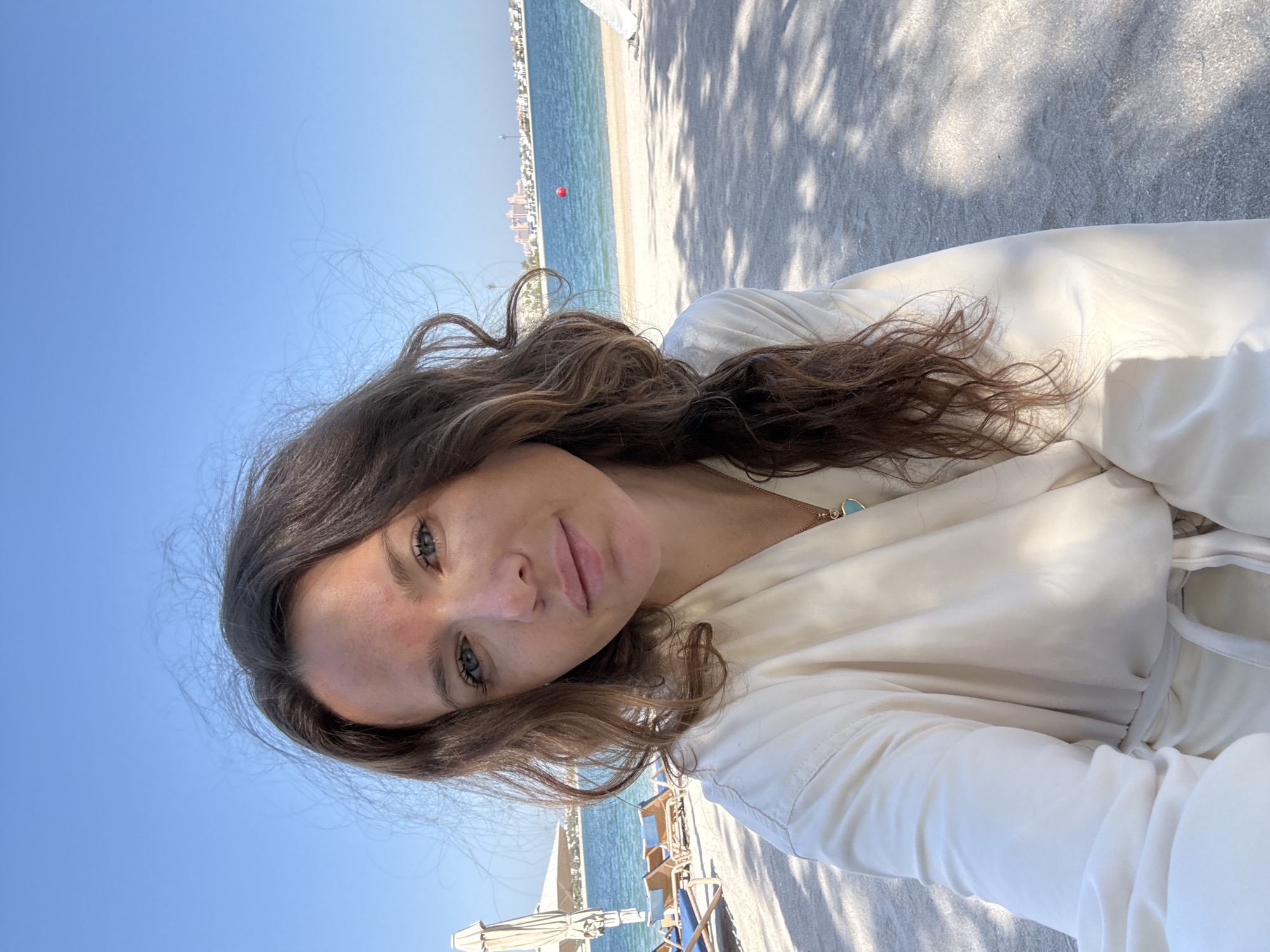 A portrait of a woman with long, wavy brown hair and blue eyes, taken on a beach in Dubai, United Arab Emirates. She is wearing a white robe and a necklace with a turquoise pendant. The background shows a beach with lounge chairs and umbrellas, the turquoise waters of the Arabian Gulf, and a distant city skyline, likely the Palm Jumeirah area, with its characteristic buildings. The sky is clear and blue.
