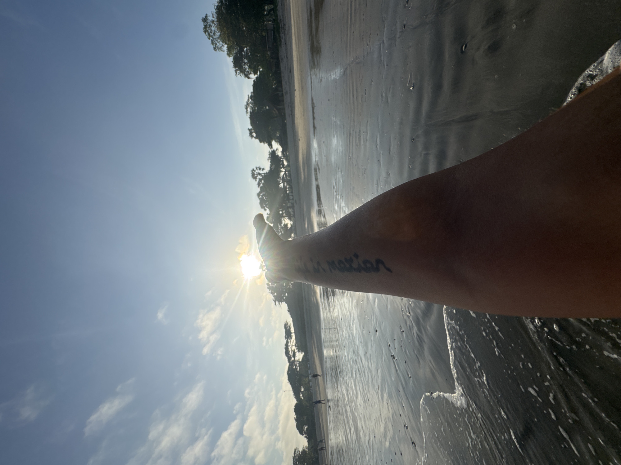The image depicts a beach scene in Jimbaran, Indonesia, during daylight hours. The sky is blue with scattered clouds. An arm is extended toward the sun, partially obscuring it, causing a lens flare. The arm has a tattoo on the inner forearm that reads, "live in motion". The beach is wet, reflecting the sky, and there are trees lining the shore in the background. Some people appear to be walking along the beach in the distance.