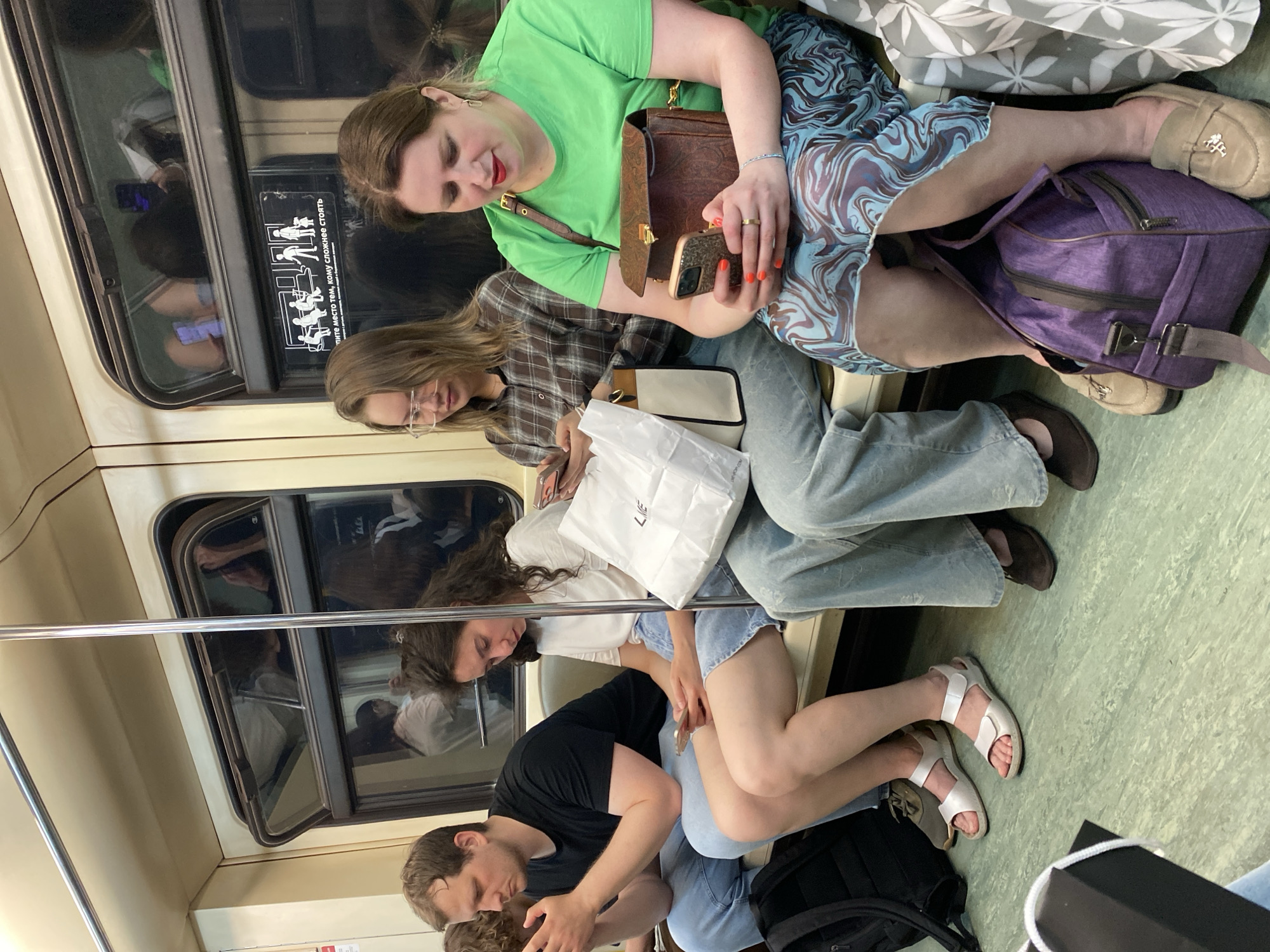 The image shows a group of people seated closely together on what appears to be a subway train. The location is Moscow, Russia, based on the design of the train car and seating.
From top to bottom:
A woman is wearing a green t-shirt and a patterned skirt. She's holding a brown handbag and using a mobile phone. Her legs are resting on top of a purple bag.
Below her, a woman in a plaid shirt is holding a white shopping bag and looking at a phone. She is wearing eyeglasses and jeans.
A woman with curly hair is wearing a white t-shirt.
At the bottom, a man in a black t-shirt is looking at a phone and appears to be leaning on the person next to him.
They are all close together in what appears to be a crowded train car. There is a metal bar visible running along the side of the car. The floors are a light green. There are windows on the left side of the car with a diagram next to one.